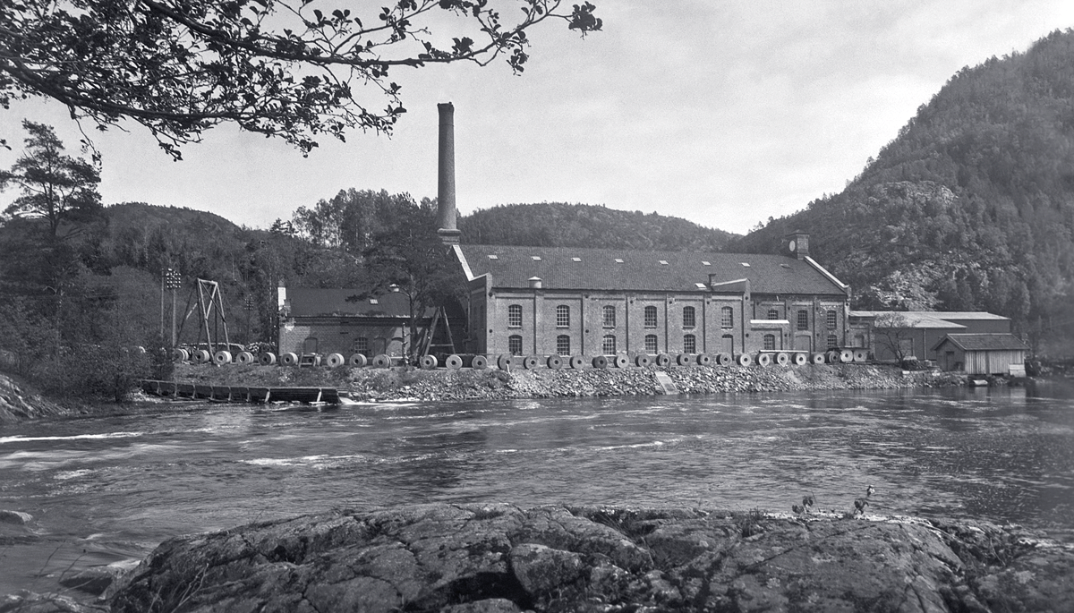 An old factory building beside a river with mountains in the background and trees in the foreground.