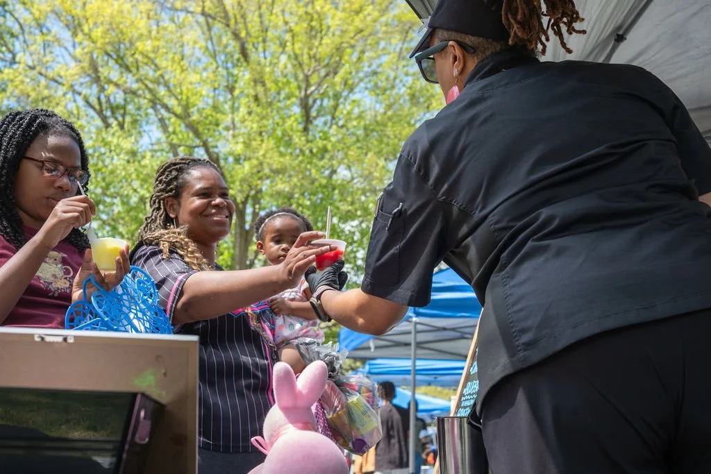 A woman in a black uniform hands a red cup with a straw to a woman with blonde dreadlocks, surrounded by children at an outdoor event under sunny skies enjoying Italian Ice.