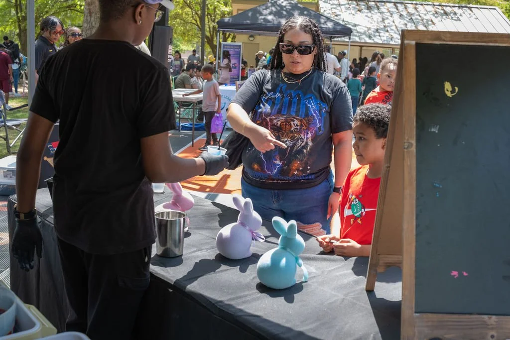 A woman and a young boy at a booth with bunny-shaped balloon figures, with other people and tents in the background at an outdoor community event serving Italian Ice.
