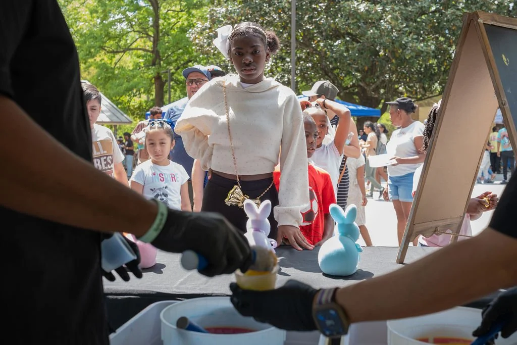 A young girl with a white bow in her hair stands at a stall with bunny-shaped items at an outdoor event, with children and adults in the background.