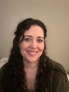 A woman with long, curly dark hair smiling in front of a plain wall.