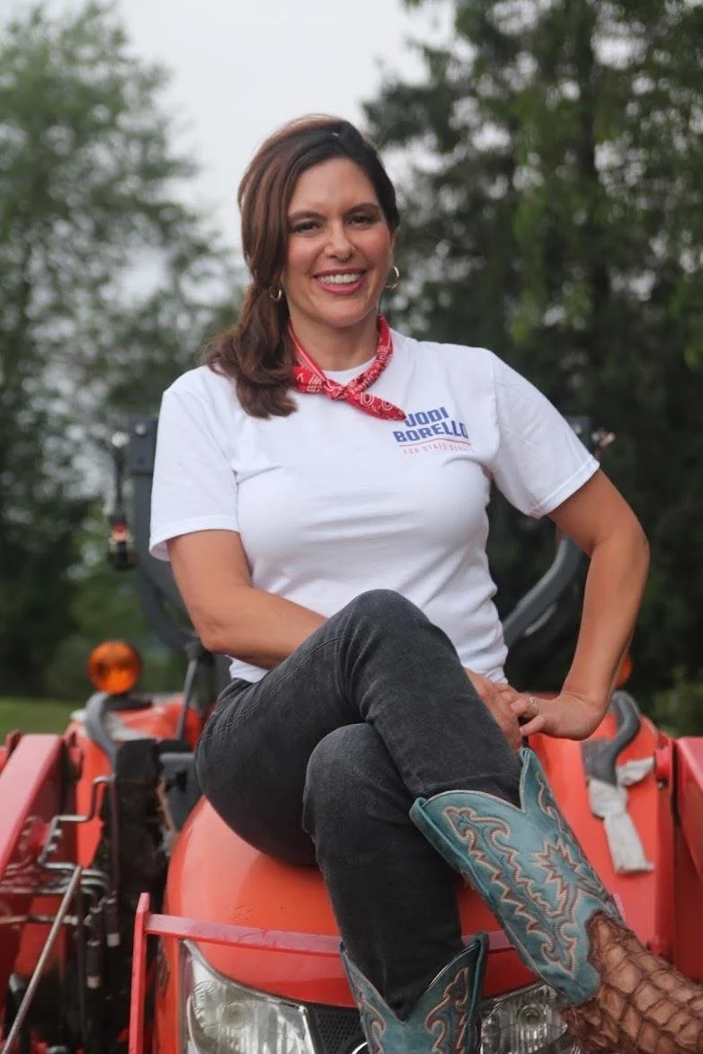 A woman sitting on a red tractor outdoors, wearing a white T-shirt, a red bandana around her neck, dark pants, and cowboy boots, smiling at the camera.