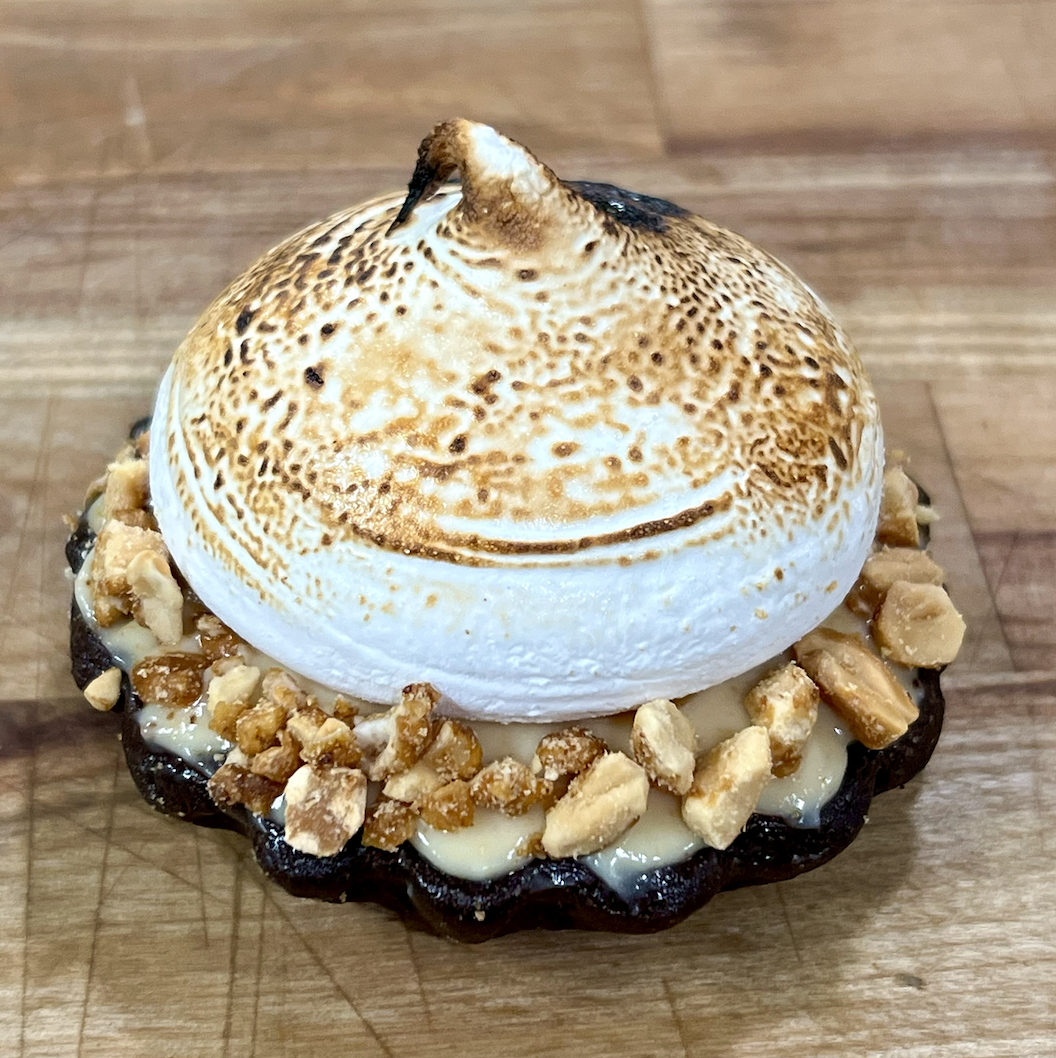 Chocolate cookie with white chocolate chips, topped with toasted marshmallow and crushed peanuts, on a wooden surface.