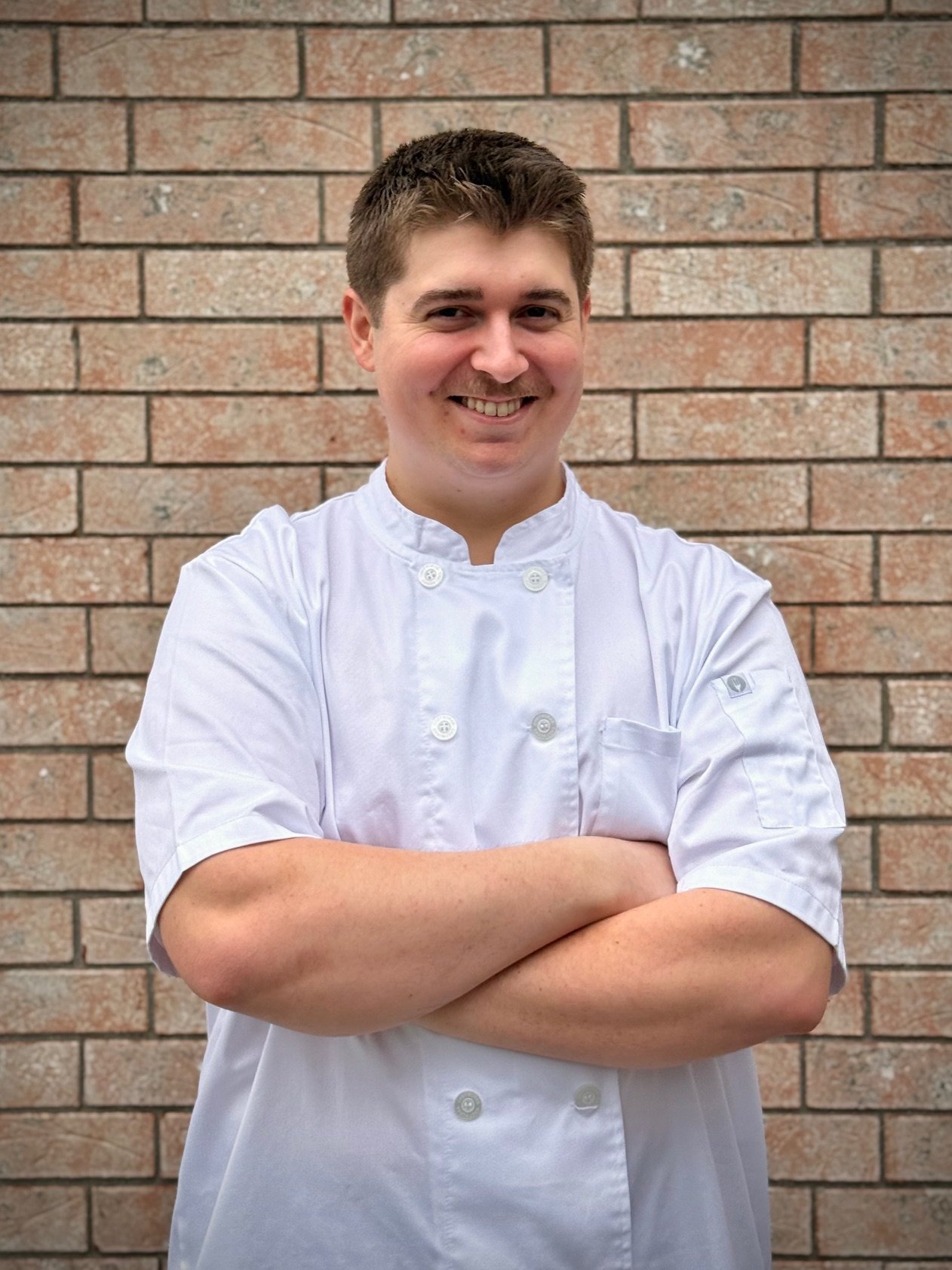 A smiling man, Chef Will, wearing a white chef's coat standing with his arms crossed in front of a brick wall.
