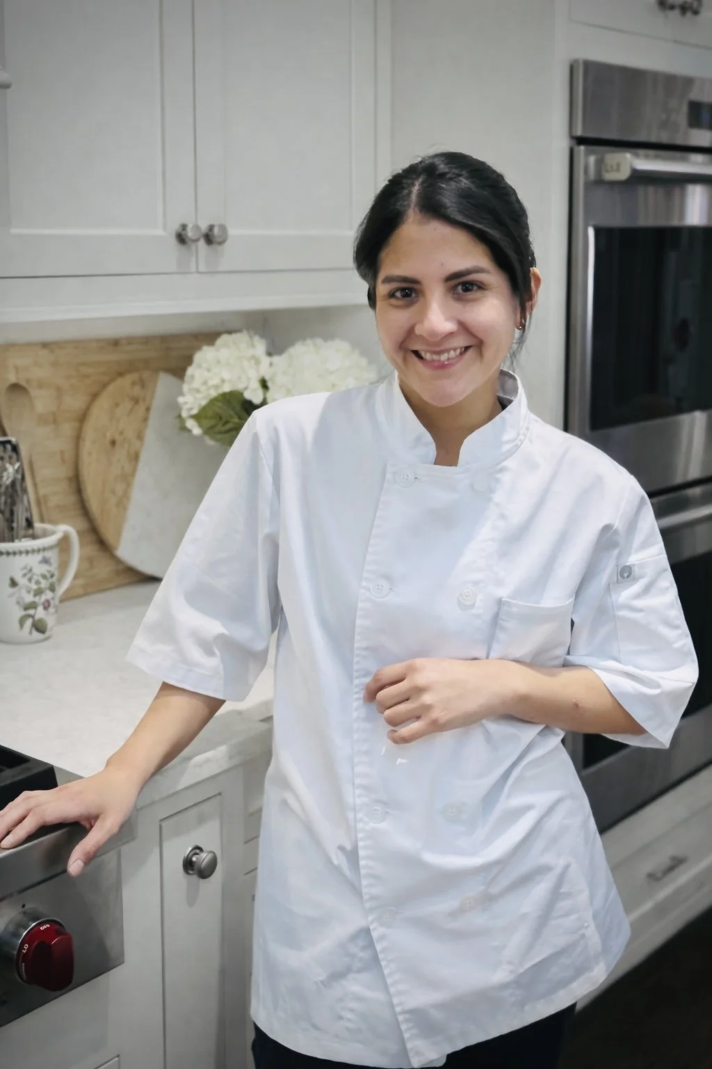 A smiling woman, Chef Magi, in a white chef's coat standing in a kitchen with white cabinetry, a vase of white flowers, and stainless steel appliances.