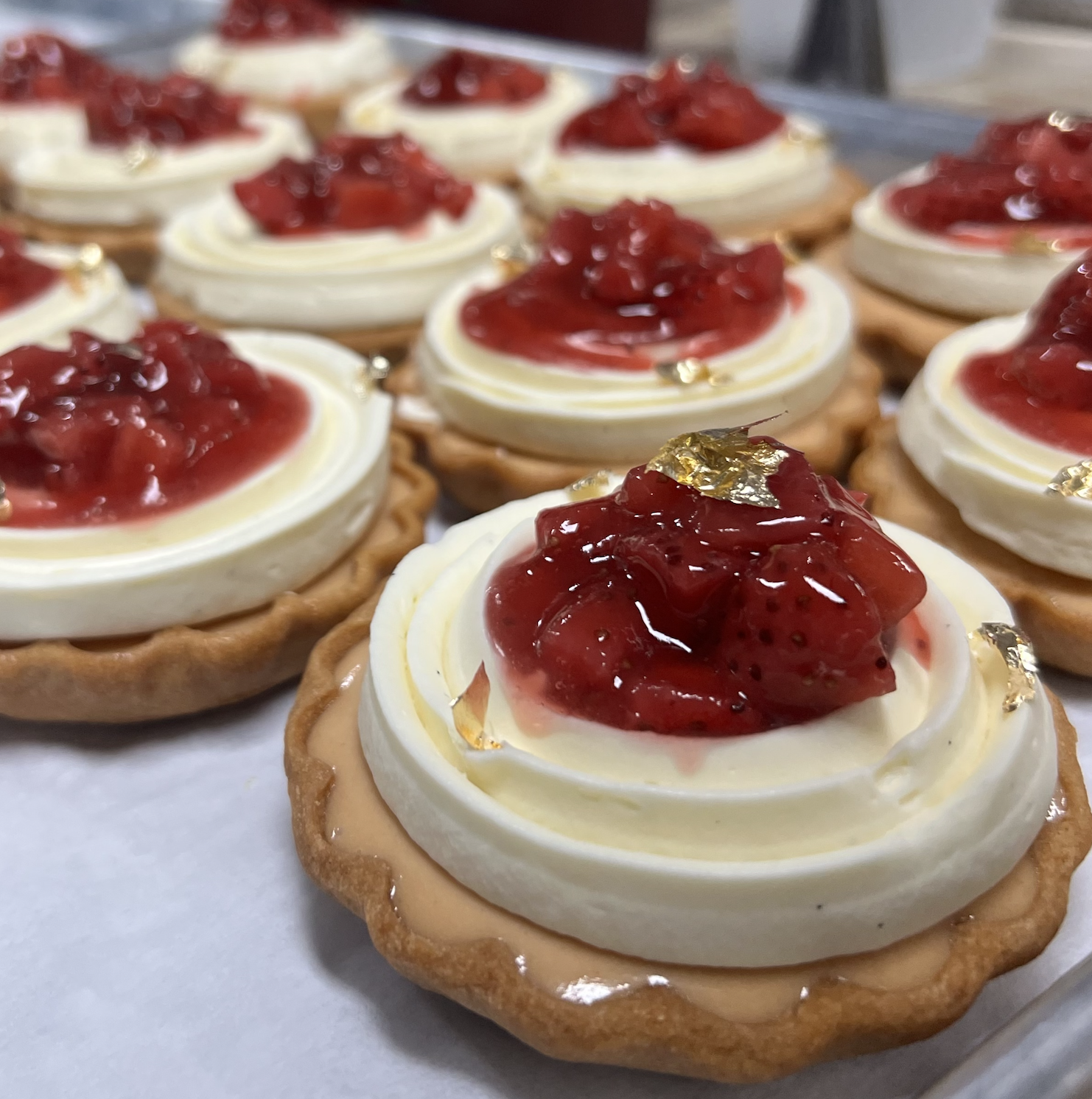Close-up of strawberry tartlets with a cookie crust, topped with whipped cream, strawberry compote, and small gold leaf decorations.