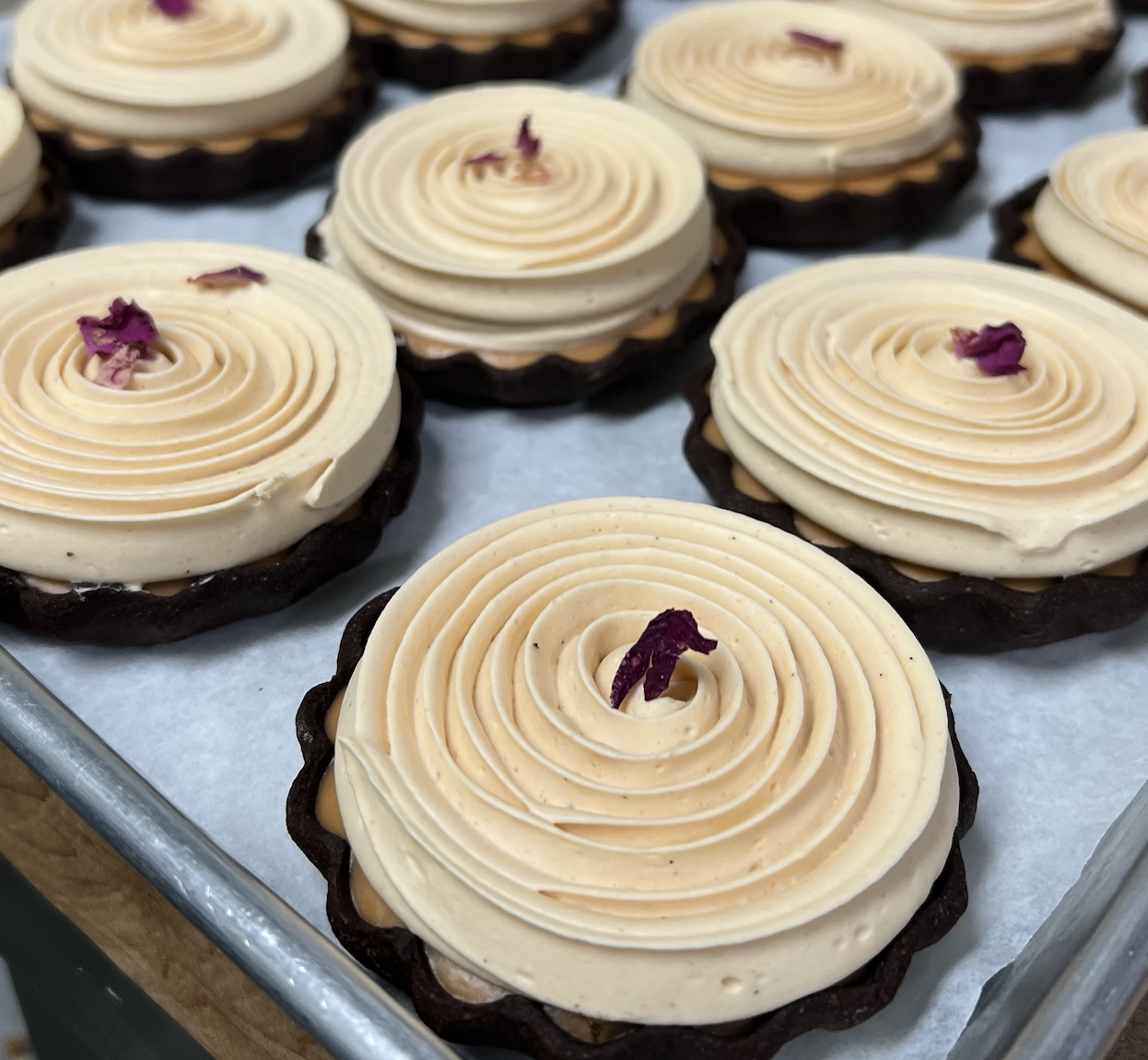 Assorted decorated tarts topped with cream and small purple flower petals on a tray.