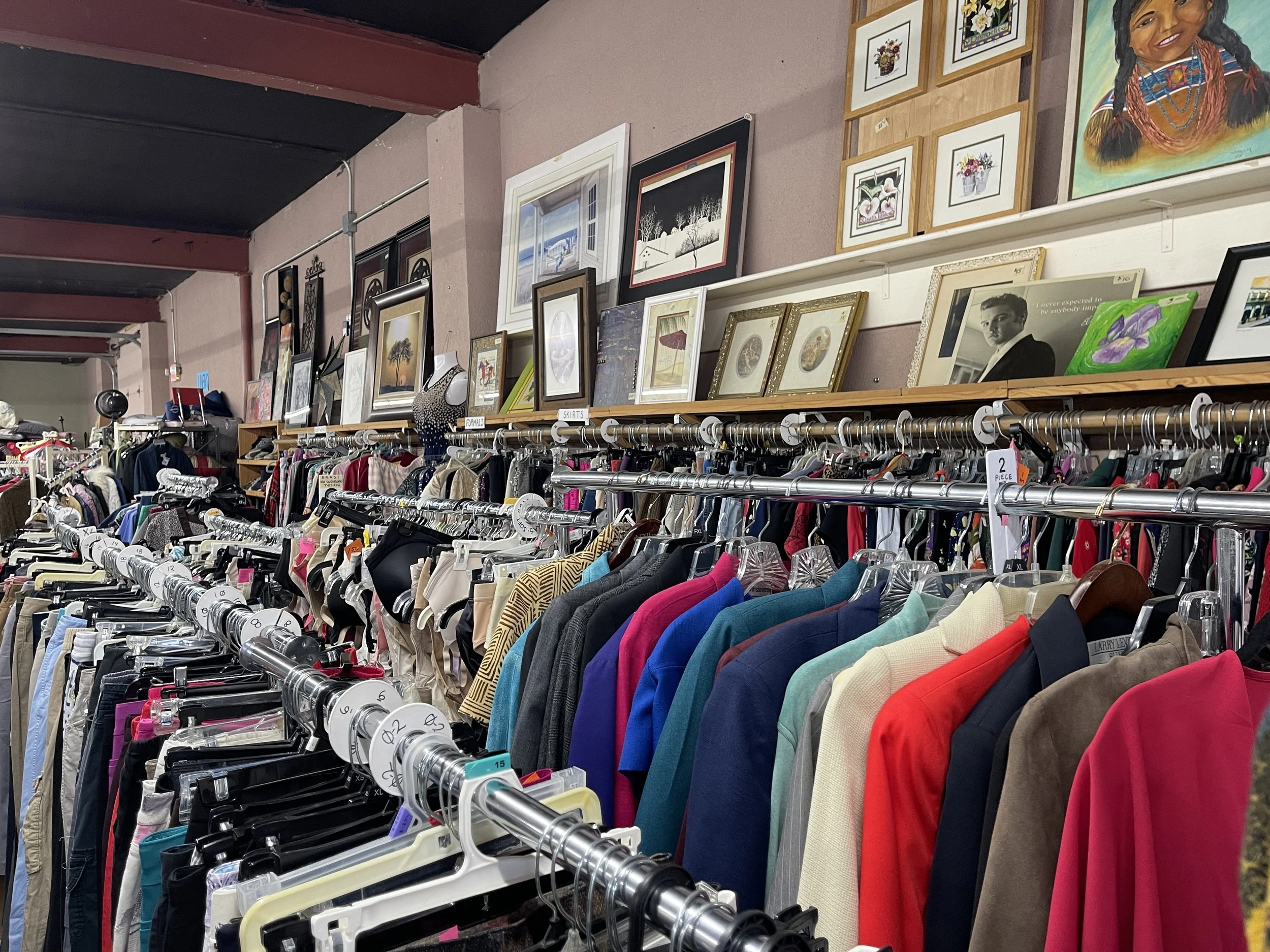 Clothing rack with various colorful clothes and framed artwork hanging on the wall above in a thrift store.