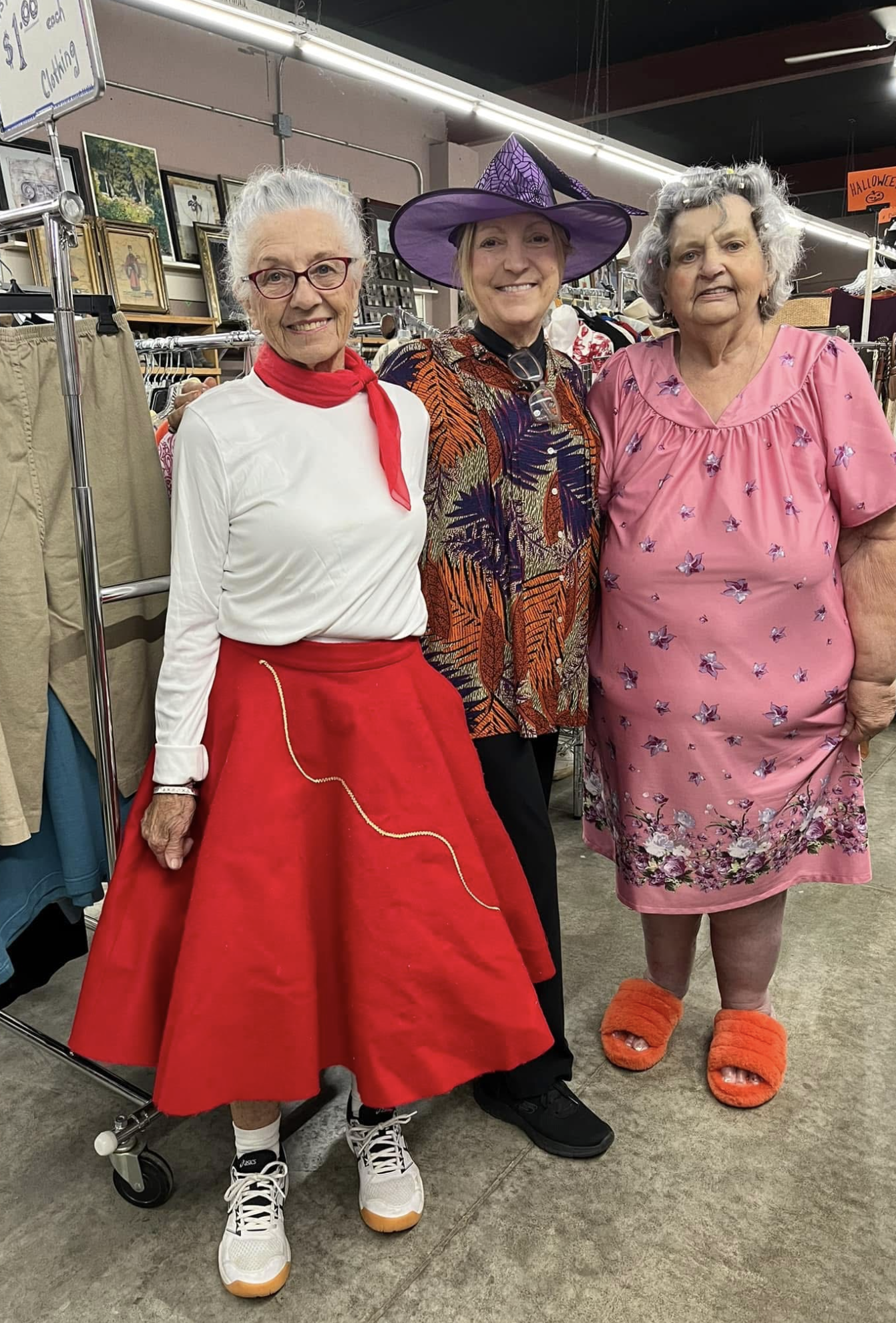 Three elderly women dressed in costumes posing together in a thrift store. The woman on the left is in a vintage outfit with a white blouse, red skirt, and a red neck scarf. The woman in the middle is dressed as a witch with a purple hat and a colorful patterned top. The woman on the right is in a pink dress with butterfly prints, wearing fluffy orange slippers.