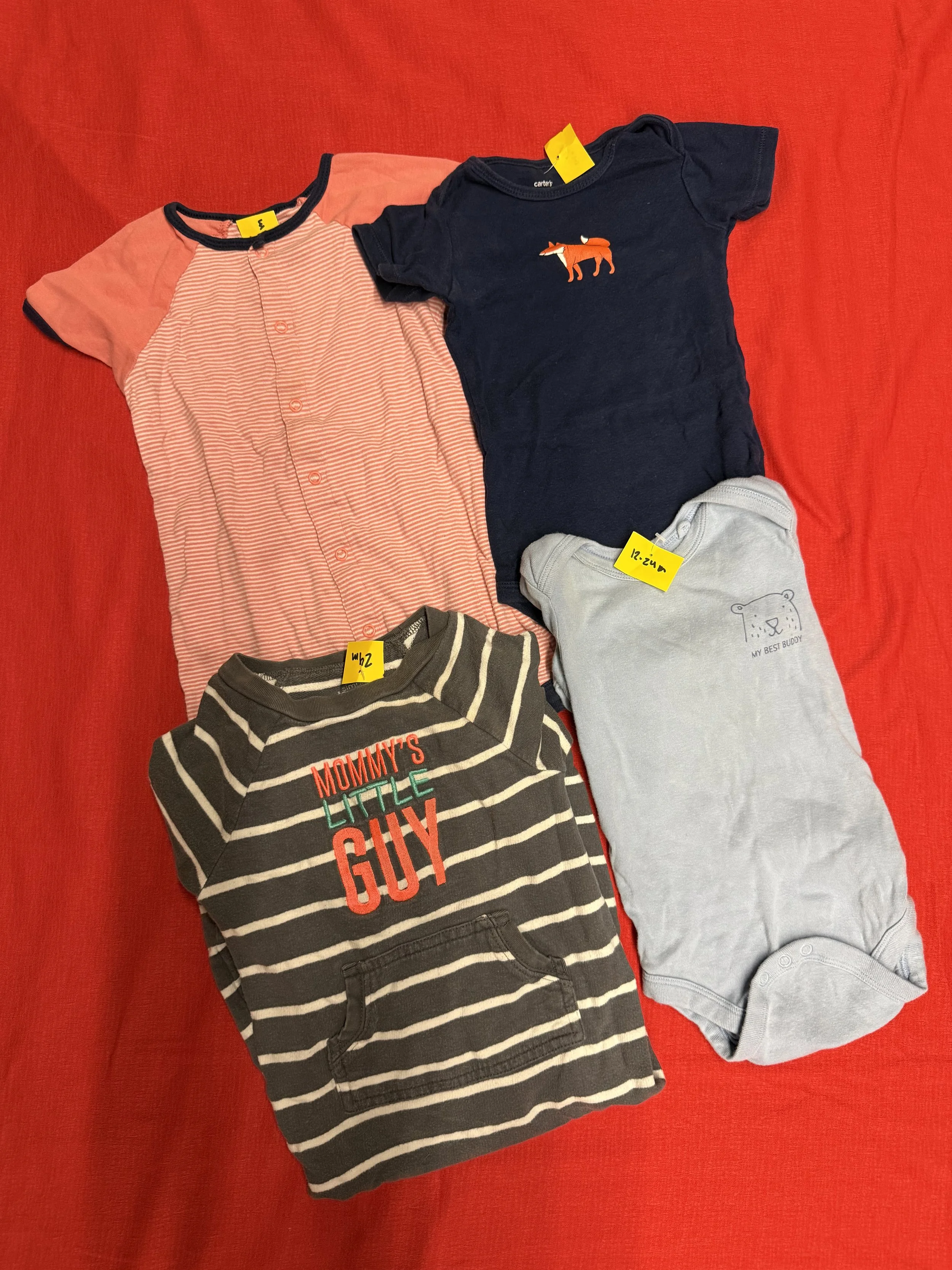 Two folded toddler clothes on a red background: a pink and navy shirt with buttons and a fox graphic, and a grey shirt with a bear graphic and a yellow price tag, alongside grey and black striped pants.