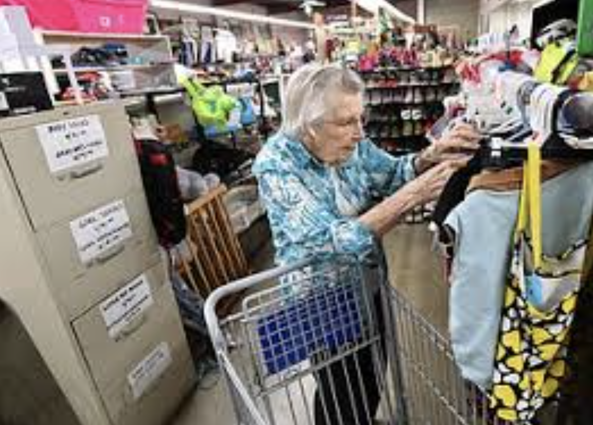 An elderly woman with gray hair wearing a blue floral shirt standing at a clothing rack in a store, holding a piece of clothing. She has a shopping cart in front of her, and there are shelves with various items like helmets and other products visible in the background.