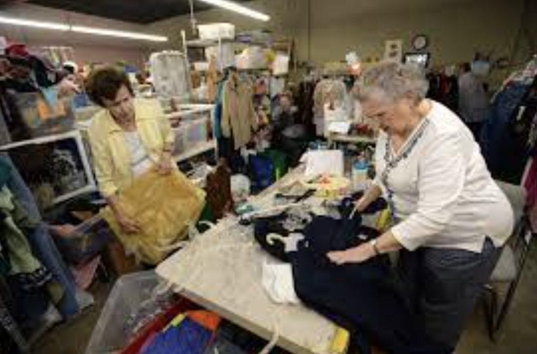 Two elderly women shopping at a thrift store or consignment shop with clothing and miscellaneous items on display.