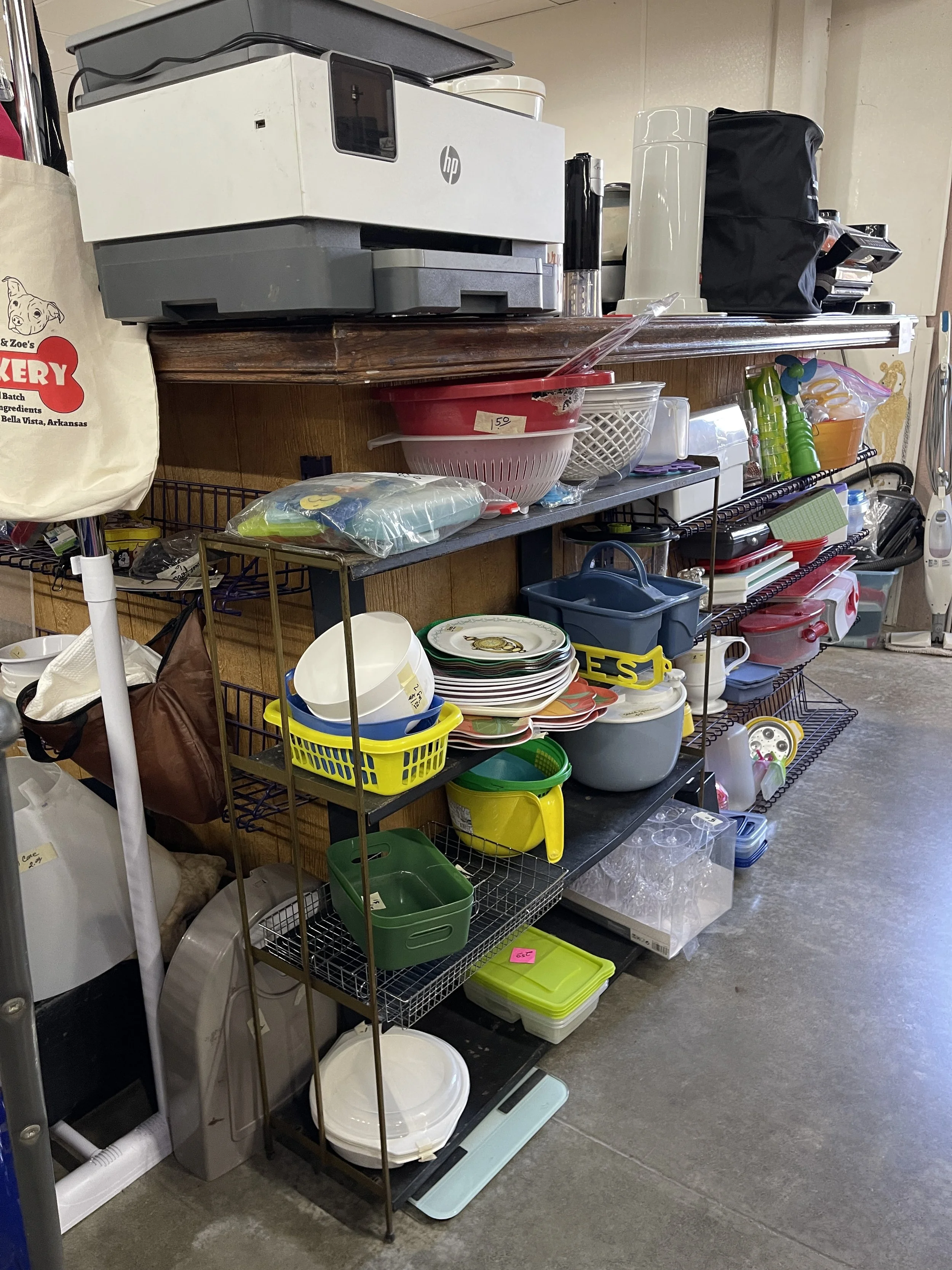 A metal shelving unit filled with various kitchen and household items, including bowls, containers, a coffee maker, and cleaning supplies. In the background, a white and gray printer or copier is on top of the shelf.