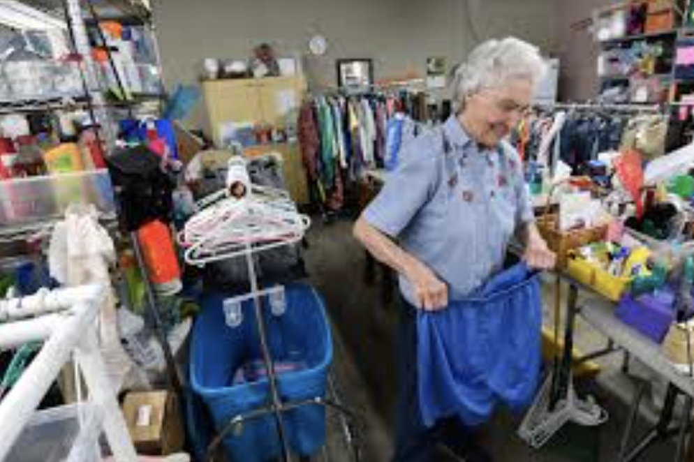 An elderly woman with gray hair in a ponytail is sorting clothes in a thrift store, surrounded by various household items and clothing on tables and racks.