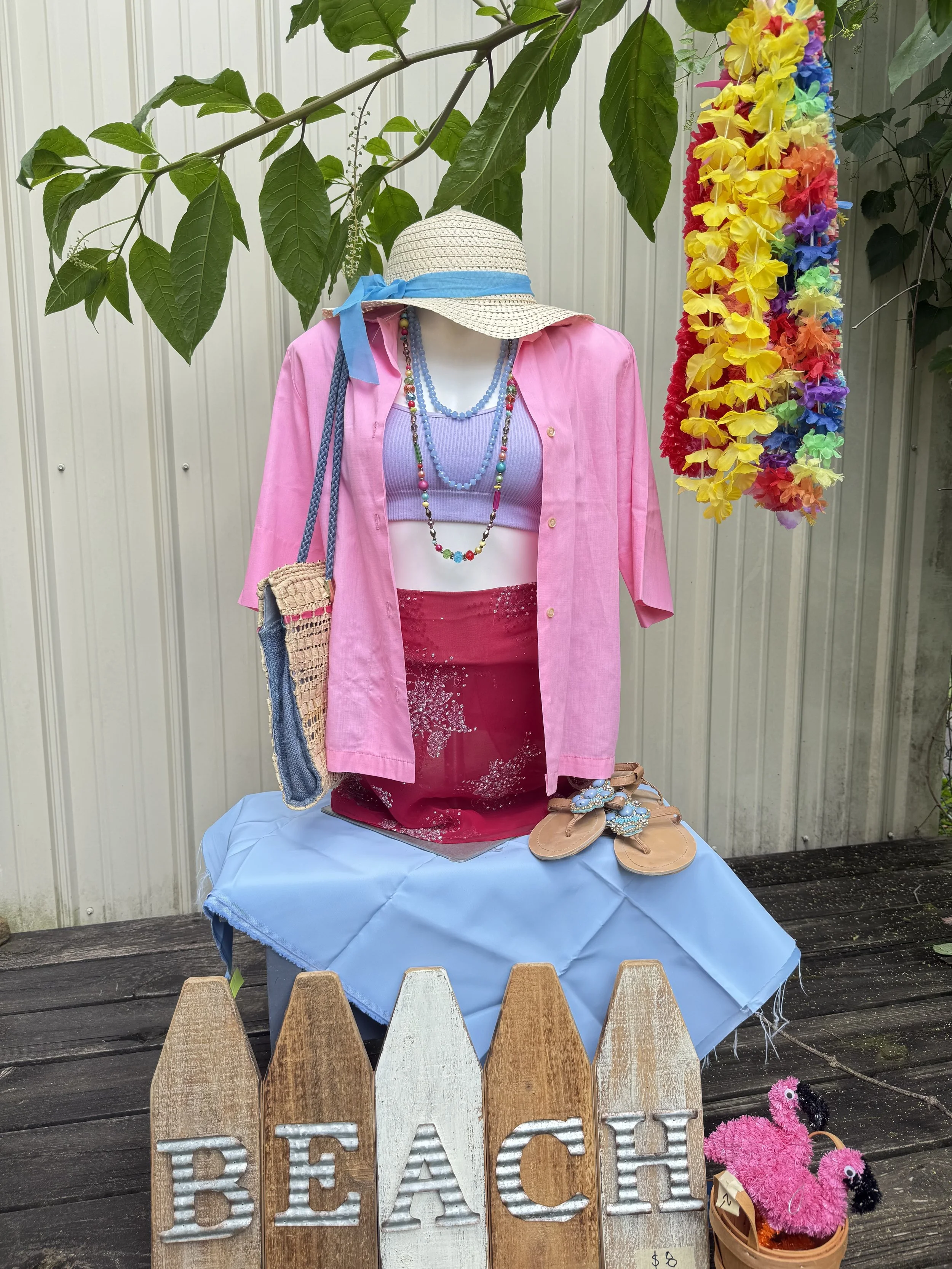 A mannequin dressed in summer clothing including a pink shirt, purple tank top, pink skirt, and multiple colorful beaded necklaces. The mannequin wears a straw sun hat with a blue ribbon. It is placed on a table covered with a blue cloth. Behind the mannequin, there is a vertical row of multicolored flower leis hanging. To the right, a basket of pink and black fuzzy plush toys is visible. In the foreground, a wooden sign spells out 'BEACH' with the letters made of corrugated metal and wood. The setting appears to be outdoors with a wooden deck and a white corrugated metal wall in the background.