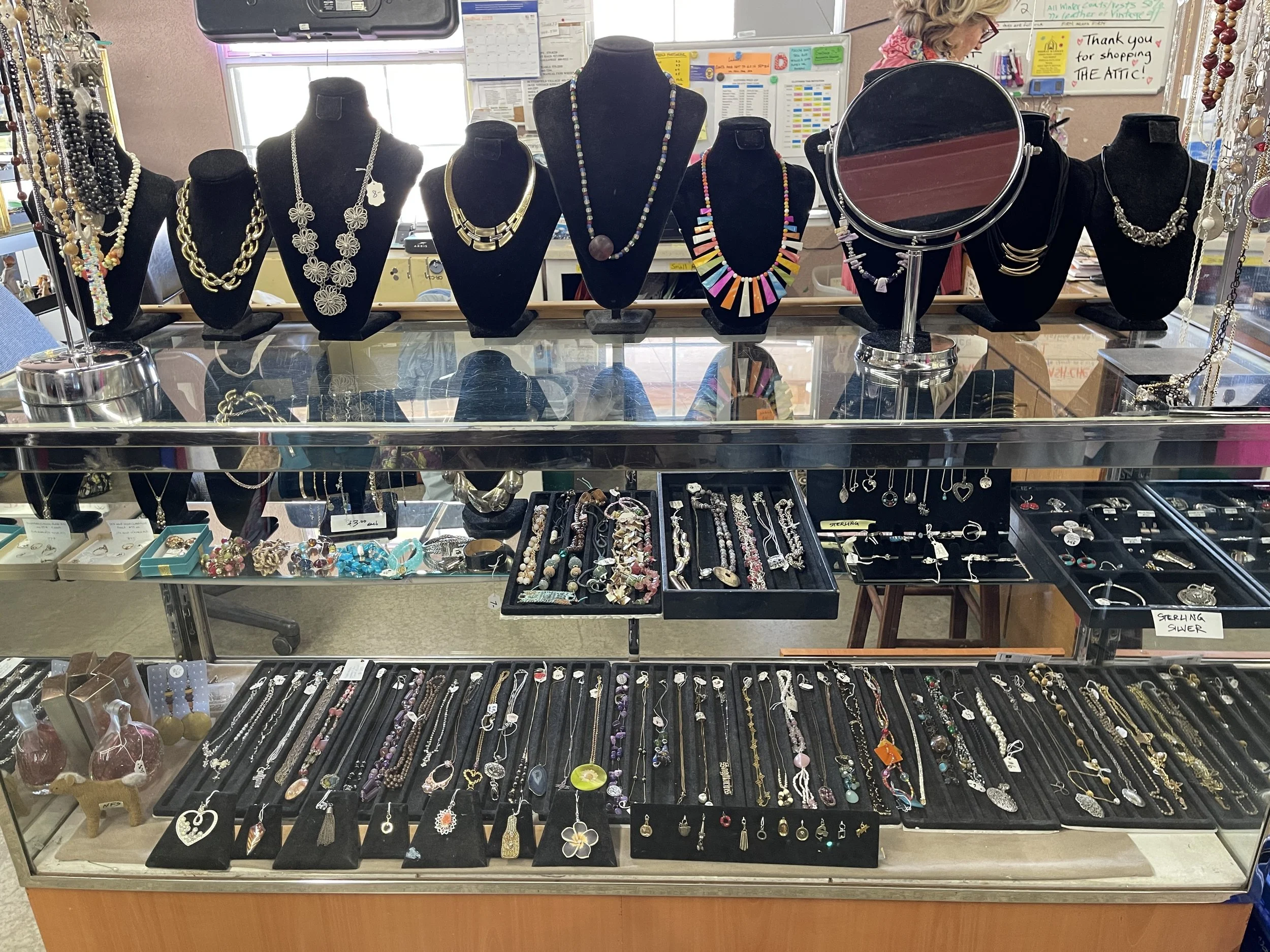 Display of various necklaces and jewelry in a jewelry store with black display busts, a tabletop with rings, bracelets, and earrings, and background items such as a mirror and shoppers.