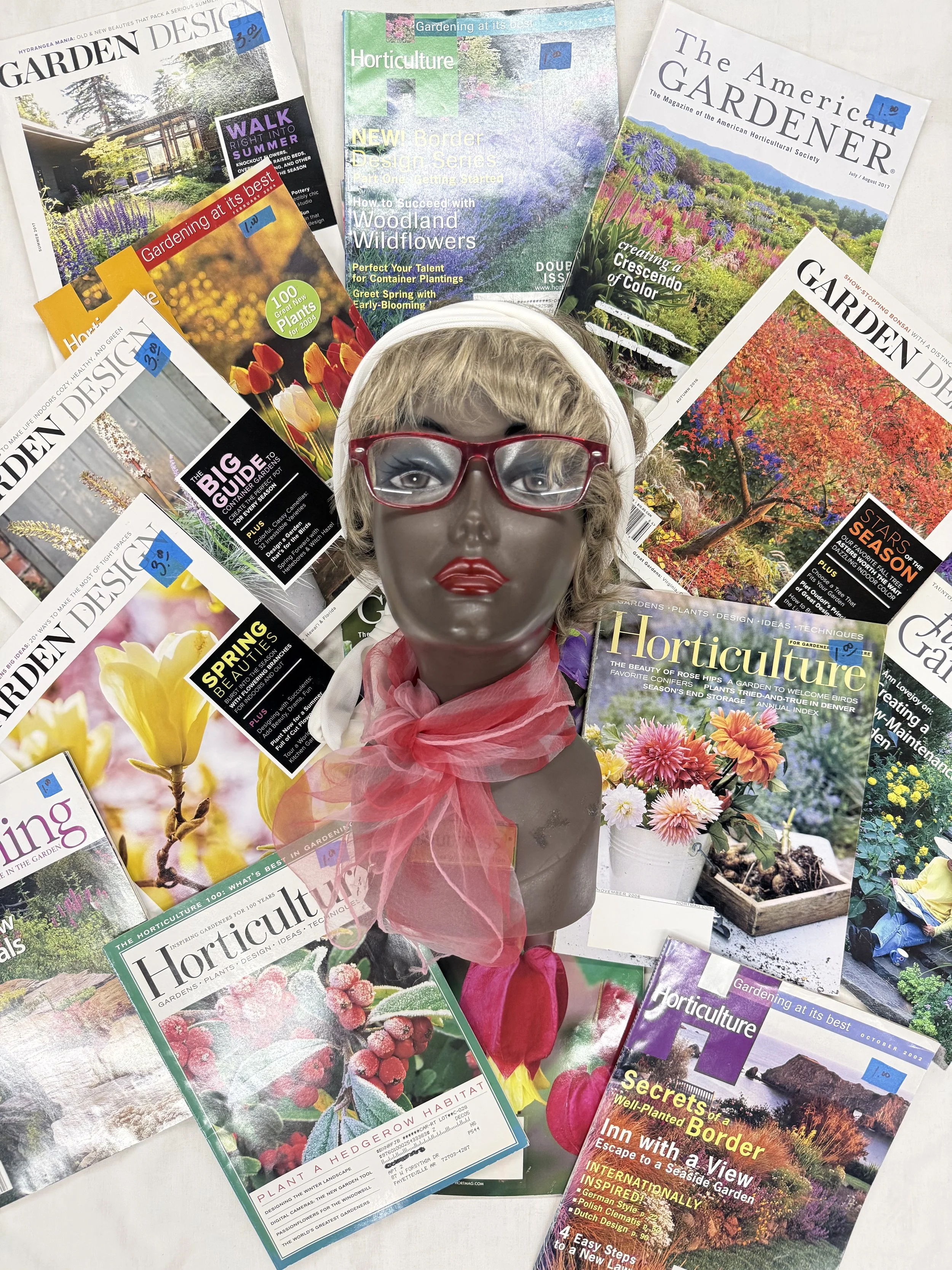 A mannequin head wearing red glasses and a pink scarf, surrounded by various magazines about gardening, horticulture, and landscape design.