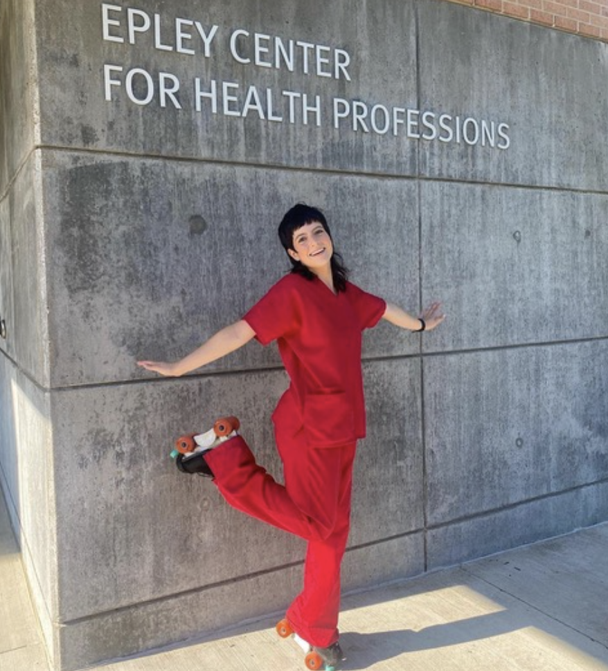 A smiling person in red scrubs standing on a sidewalk next to a concrete wall with the sign 'E P L E Y CENTER FOR HEALTH PROFESSIONS' mounted on it, with one leg raised behind them on a roller skate.