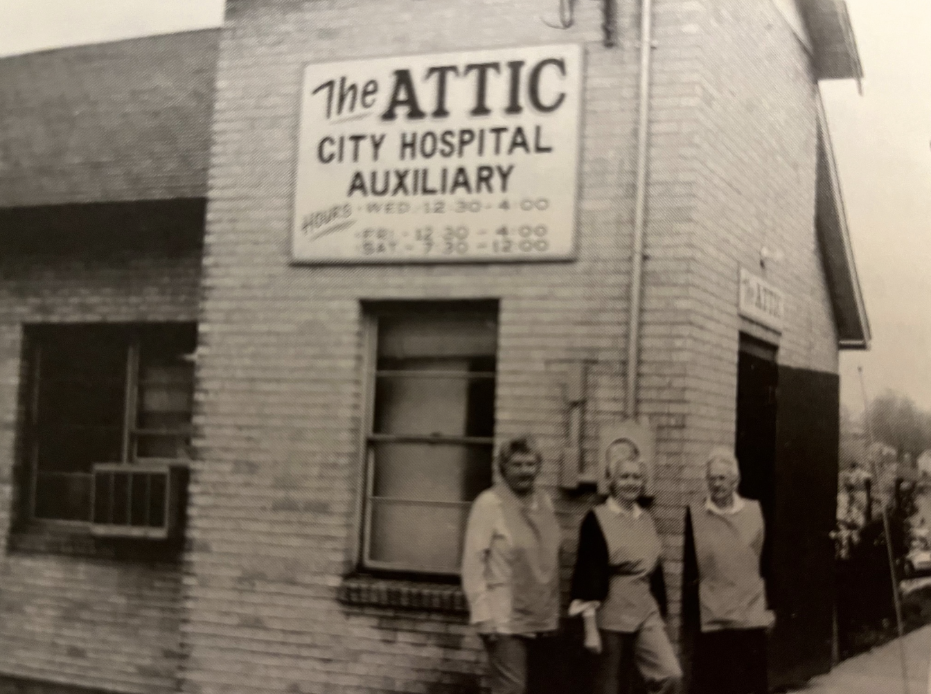 Black-and-white photo of three people standing in front of a brick building with a sign that reads "The Attic City Hospital Auxiliary."