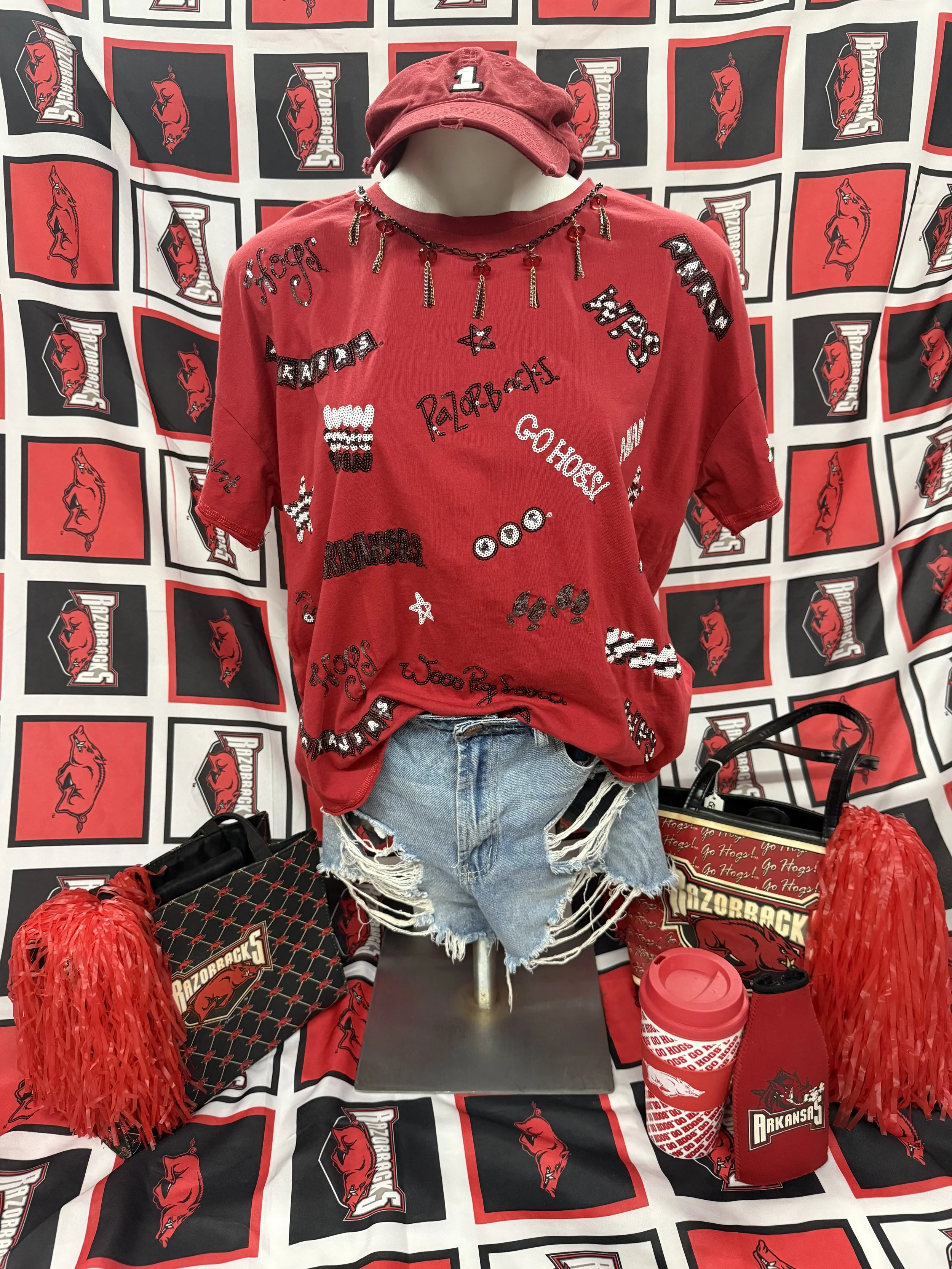 Display of Arkansas Razorbacks apparel and accessories on a mannequin, including a red t-shirt with various Razorbacks logos and phrases, a red baseball cap with the number 1, a black beaded necklace with charms, distressed denim shorts, a black bag with Arkansas Razorbacks logo, striped red and white reusable cup, red pom-poms, and a Razorbacks-themed shopping bag, set against a background with multiple Razorbacks logos.