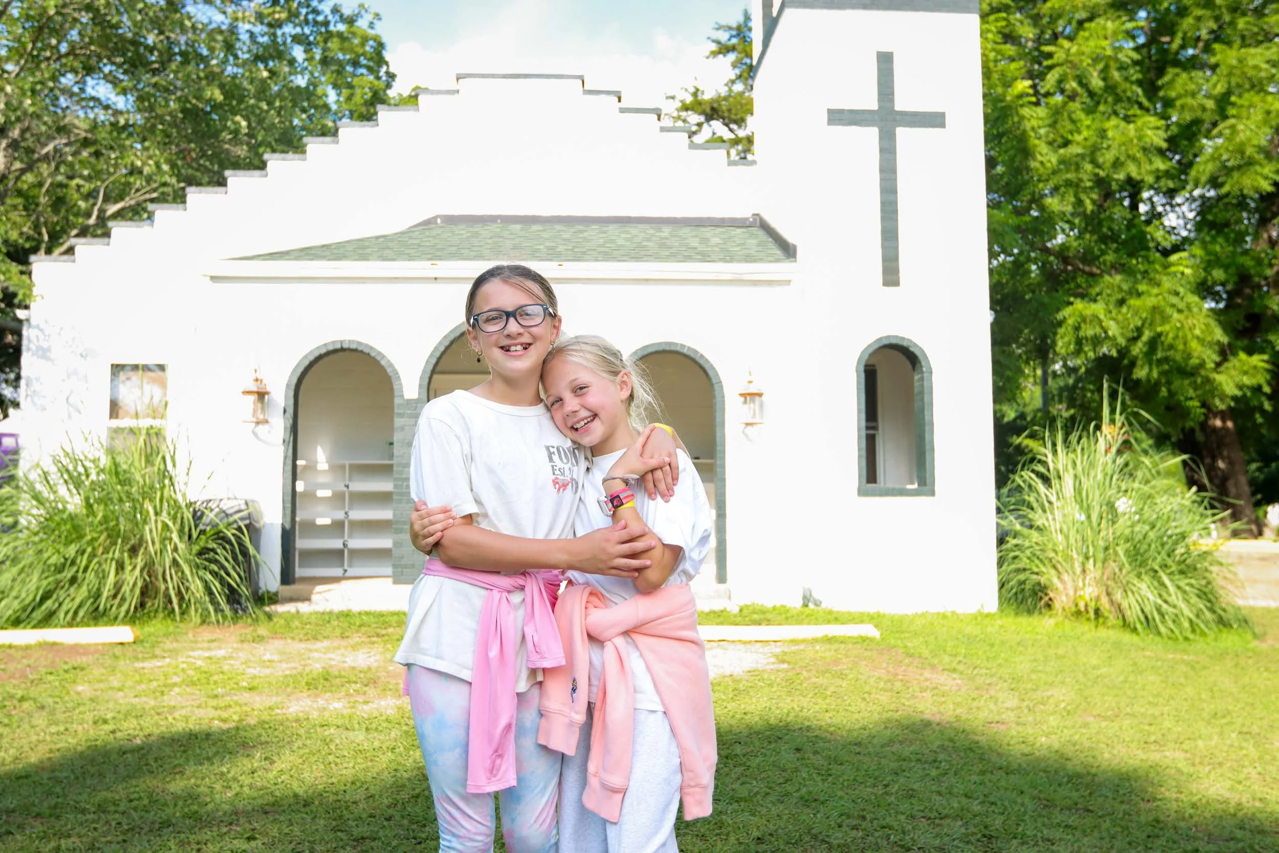 Two smiling young girls hugging in front of a chapel.