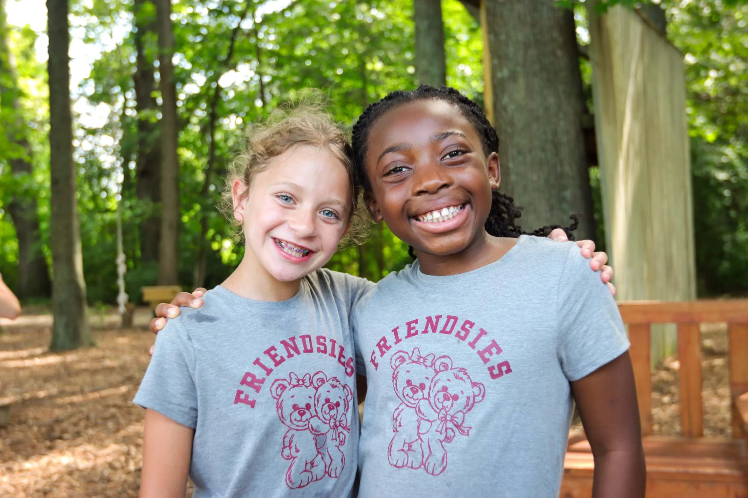 Two girls wearing matching shirts smiling and hugging in a wooded outdoor area.