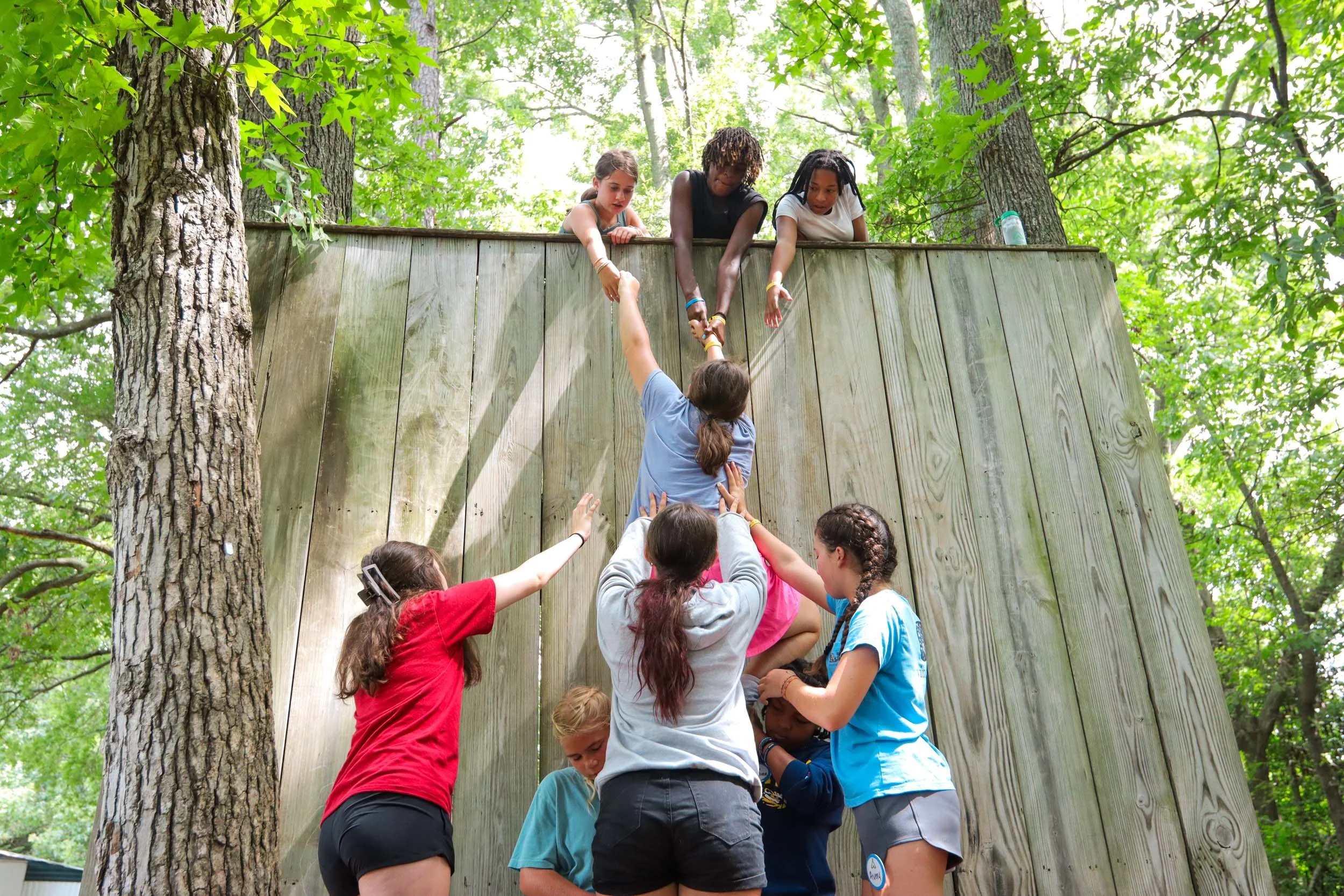 Camper team-building activity where the children are helping to lift a girl up a wooden wall.