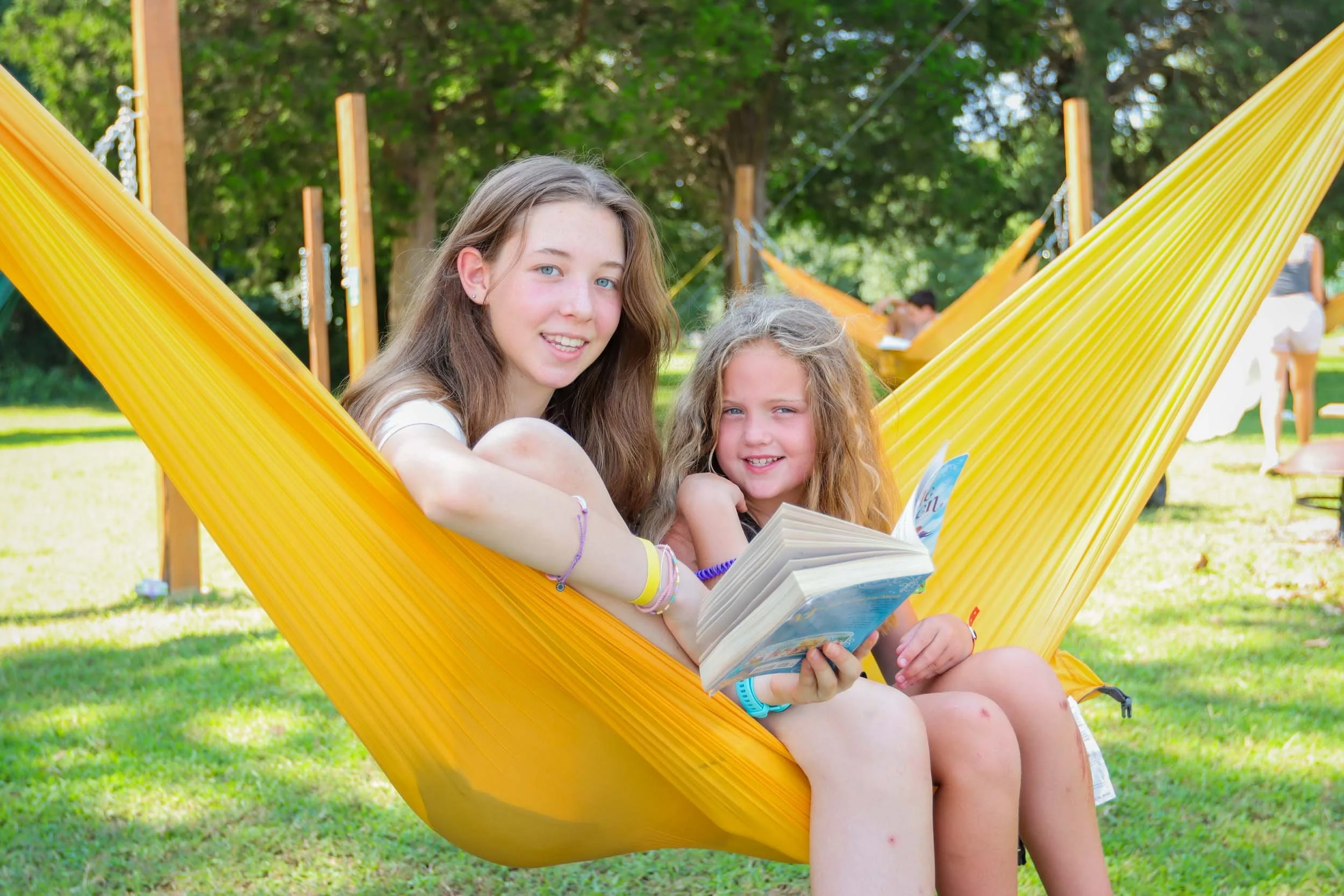 Two girls, a counselor and camper sitting in an outdoor yellow hammock reading a book.