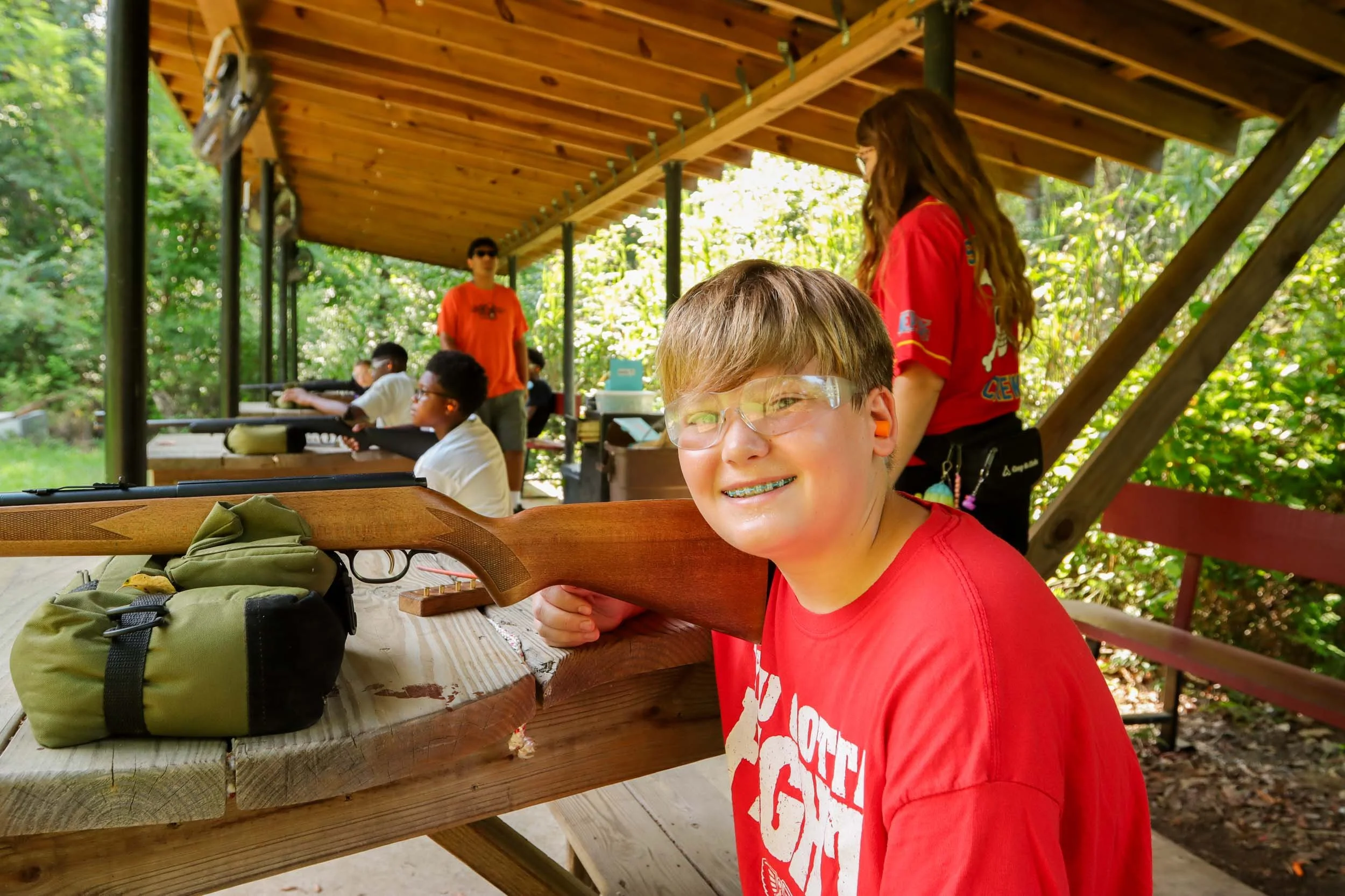A young boy smiling and holding a rifle at an outdoor shooting range.