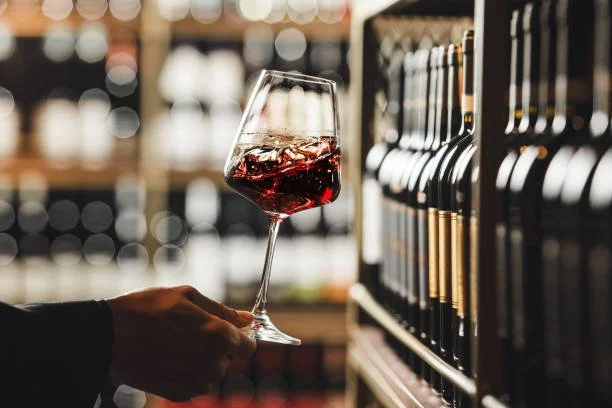 A person holding a glass of red wine in front of wine bottles on a store shelf.