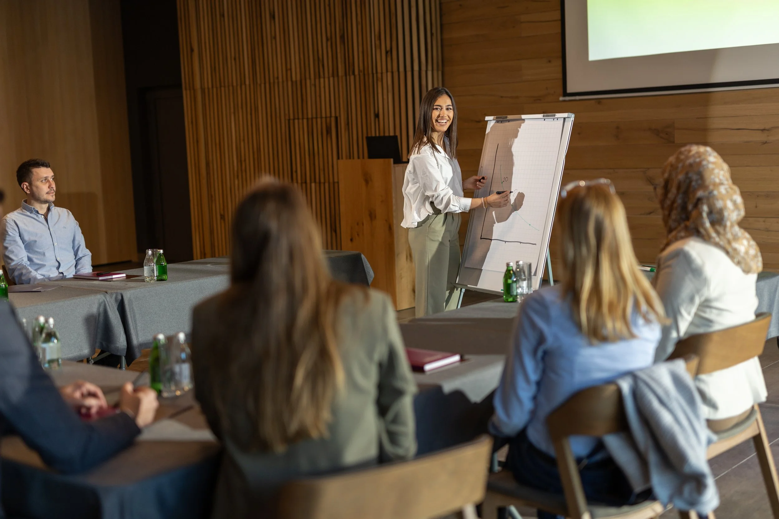 A woman is giving a presentation to a group of people in a conference room, standing next to an easel with papers and a large screen behind her.