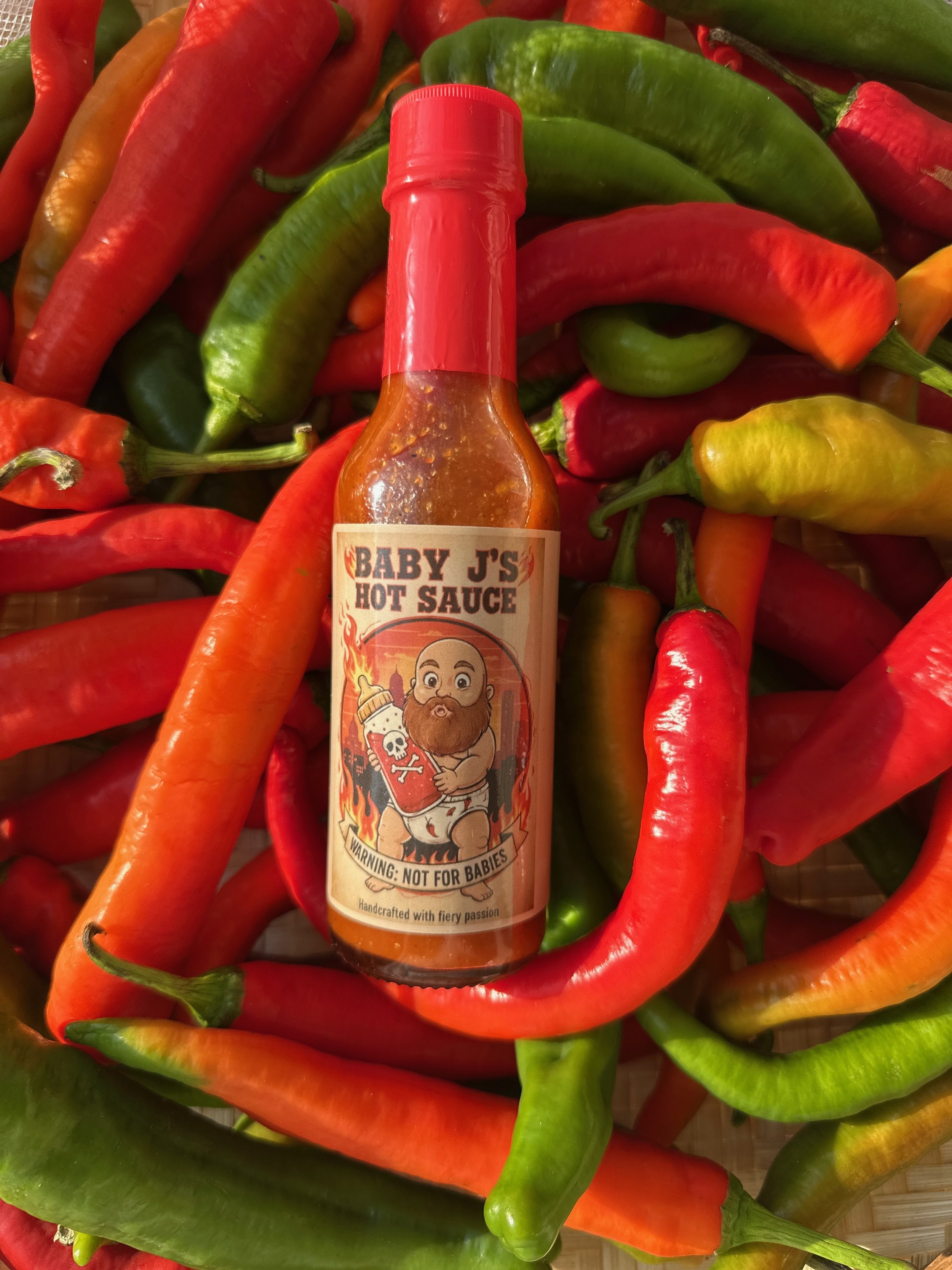 Bottle of hot sauce labeled 'Baby J's Hot Sauce' with a cartoon baby holding a bottle of hot sauce, surrounded by colorful red, green, and yellow chili peppers.