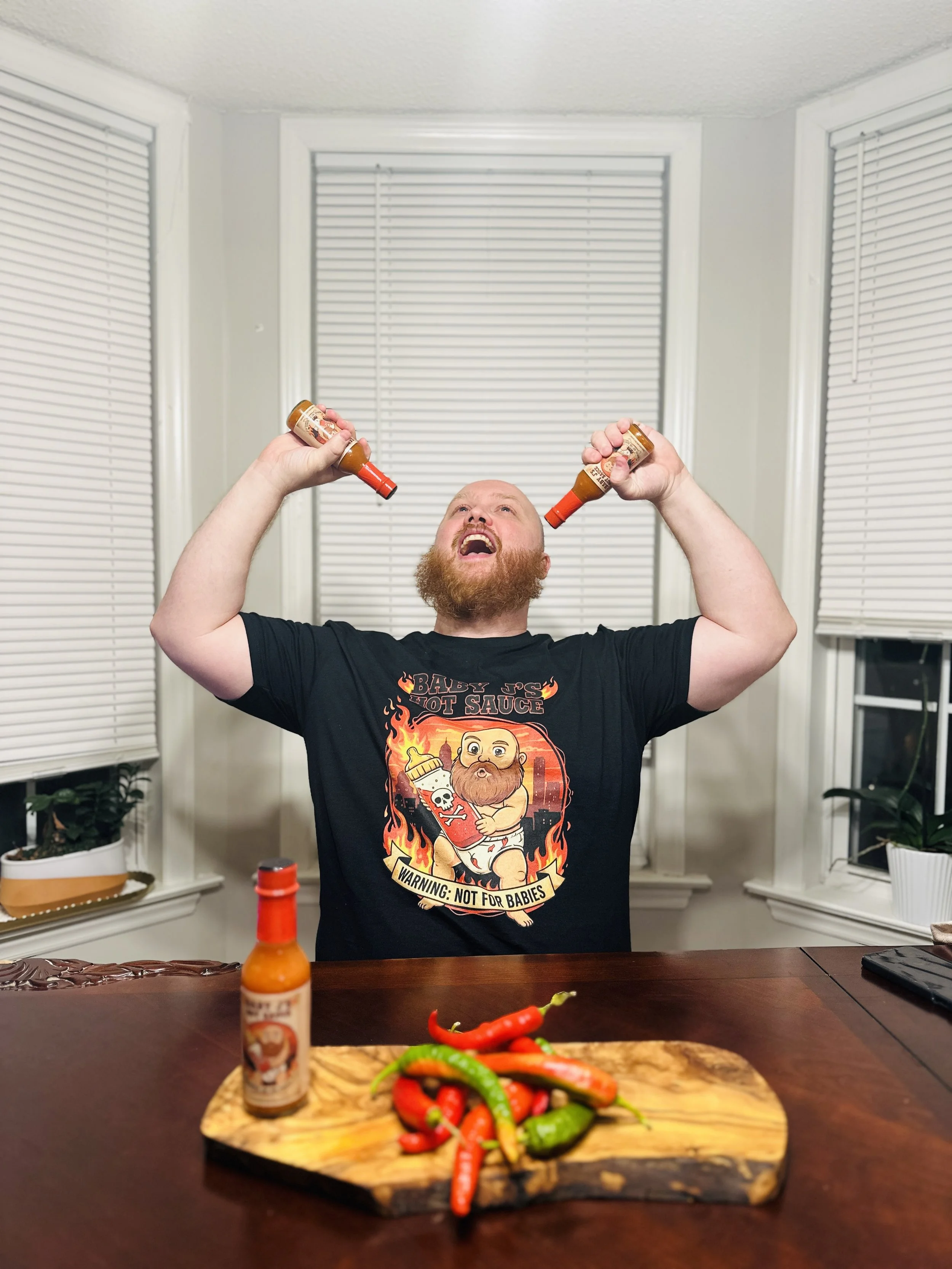 A man with a red beard and wearing a black T-shirt with a graphic design of a baby holding hot sauce bottles sits at a table with a wooden cutting board and a variety of colorful chili peppers. He is holding two bottles of hot sauce, one in each hand, and looking up with an excited expression.