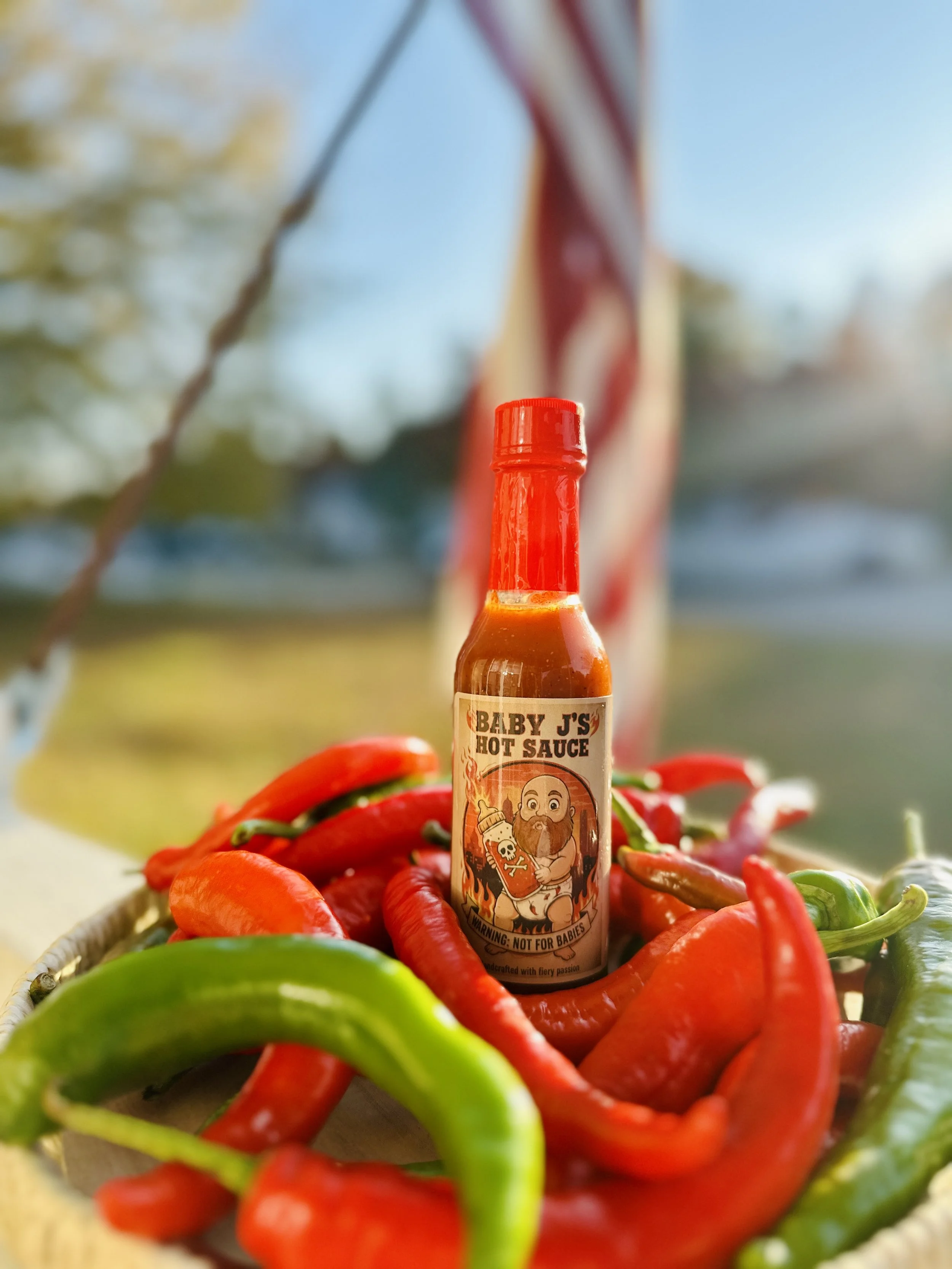 Bottle of Baby J's hot sauce with red and green chili peppers in a basket outdoors.