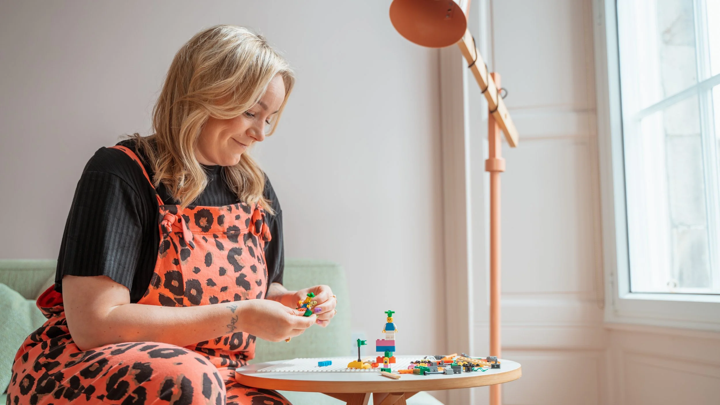 A woman wearing a black shirt and a red leopard print apron, sitting on a green couch, building with LEGO bricks on a small round table. There are LEGO creations and pieces on the table, a large window with white trimmings behind her, and a tall orange floor lamp beside her.