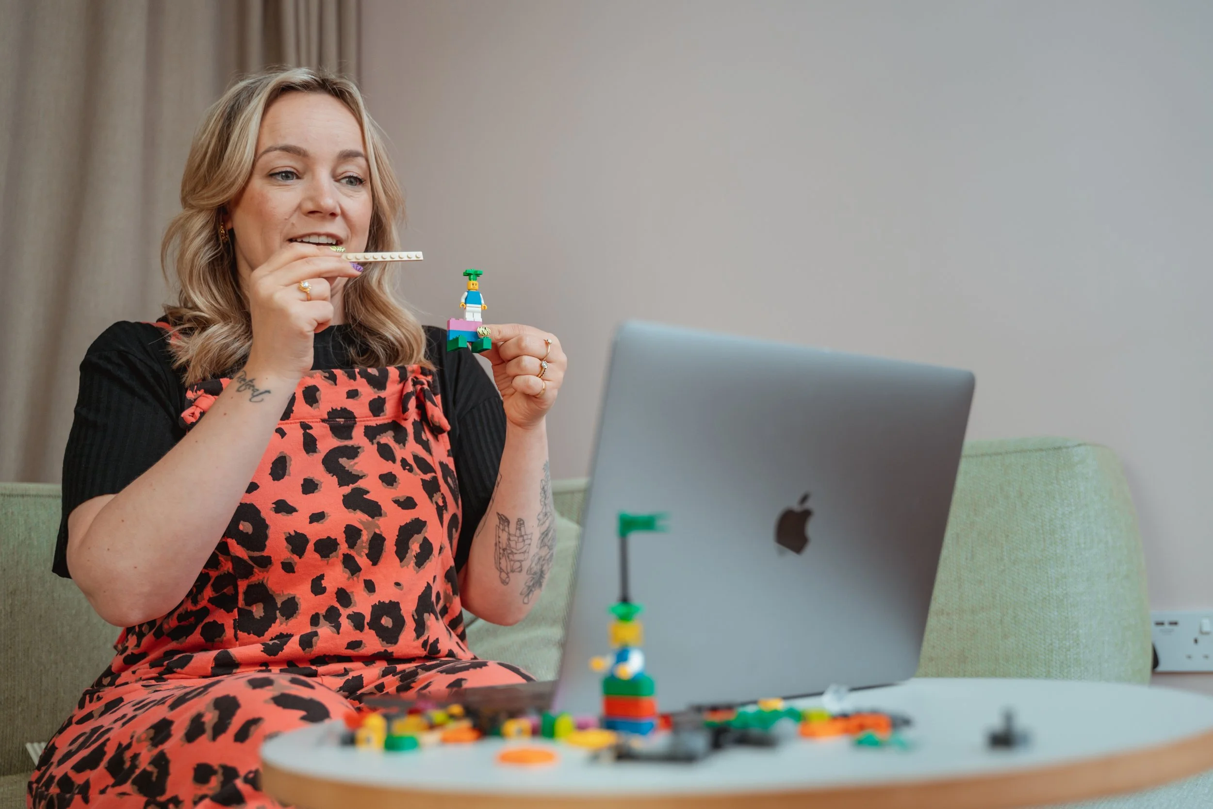 Woman sitting on a green couch assembling a small LEGO figurine while on a laptop, with LEGO pieces spread on a white table.