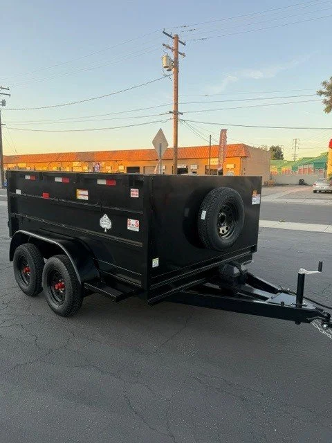 Black utility trailer with dual axles and a spare tire, parked on a street with power lines and buildings in the background during sunset.