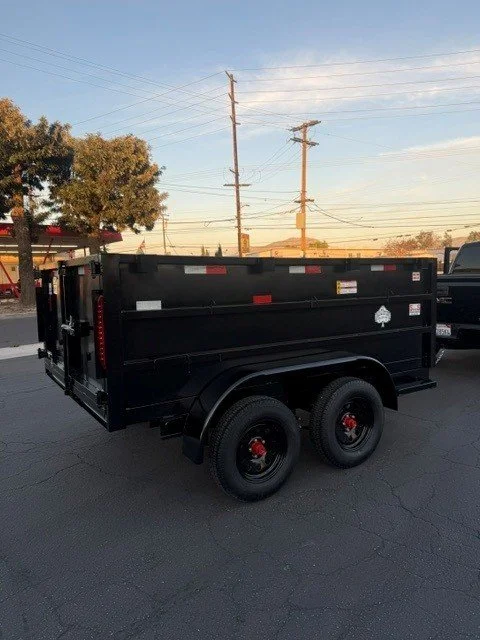 Black utility trailer with dual axles parked on pavement, with power lines, trees, and a building in the background during sunset.