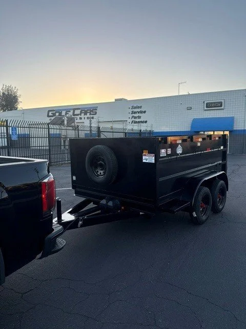 A black utility trailer attached to a pickup truck parked in a lot near a fenced area with a large building in the background that has signage for car sales, service, parts, and finance.