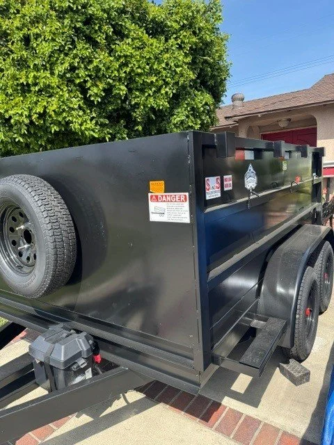 Black utility trailer with a spare tire mounted on the front, parked on a concrete driveway next to a red house with a brown shingle roof, partly obscured by green bushes.