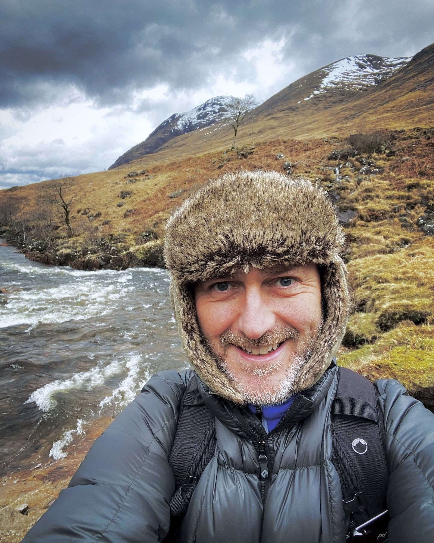 A man outdoors near a river, wearing a furry hat, a black jacket, and a backpack, smiling at the camera with mountains and cloudy sky in the background.