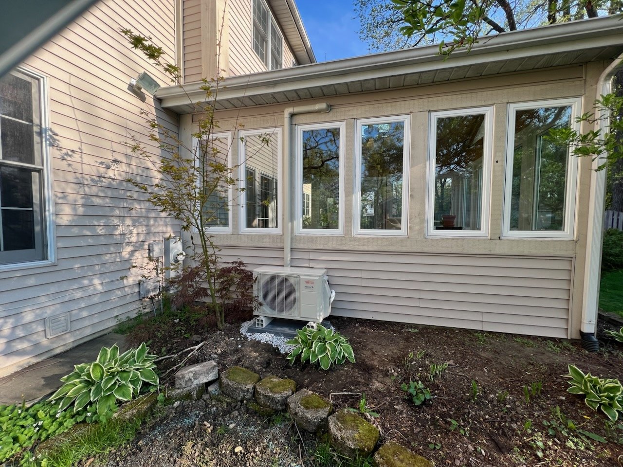 Backyard garden area of a house with a small tree, several green and variegated plants, and an air conditioning unit. The house has beige siding and multiple windows, with some trees and blue sky visible in the reflection.