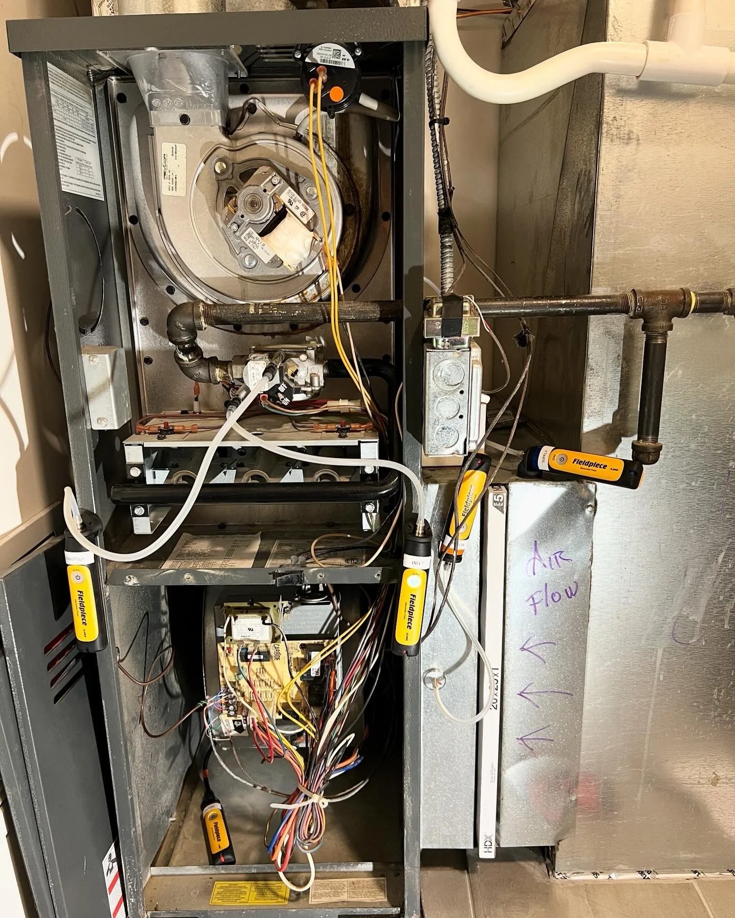 Inside view of an HVAC furnace showing various components, wires, and pipes, with a handwritten note on a metal panel indicating airflow direction.