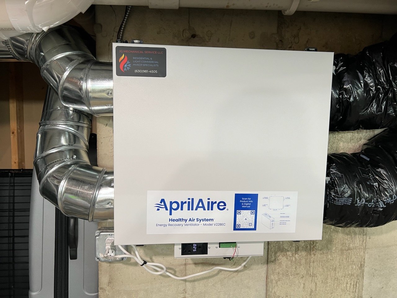 An Aprilaire energy recovery ventilator system installed on a wall, with metallic ductwork connected to it for ventilation and airflow. The system has a label indicating it is a healthy air system.
