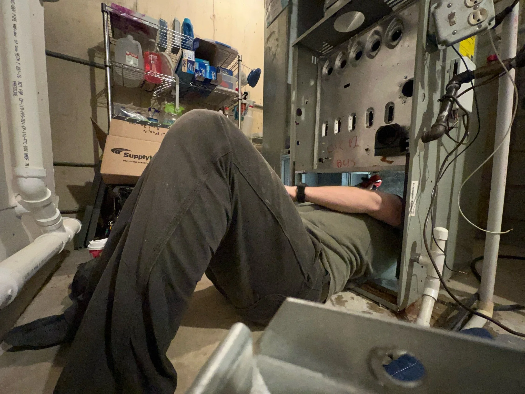 A man lies on his back under a large electrical or mechanical panel, working on it with tools in hand in a basement or utility room with pipes and shelves with supplies.