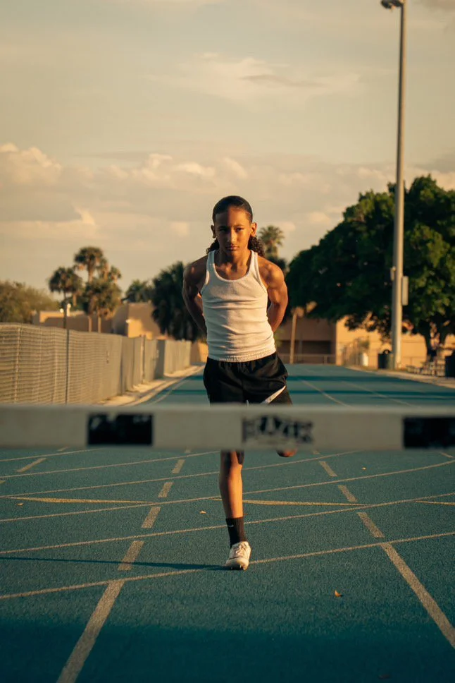 A young athlete running on an outdoor track and crossing the finish line, wearing a white tank top and black shorts during sunset.