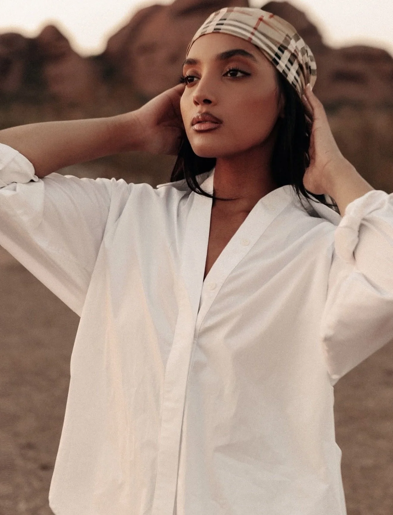 Young woman with dark hair wearing a white shirt and a patterned headscarf, standing outdoors with a rocky background at sunset or sunrise.