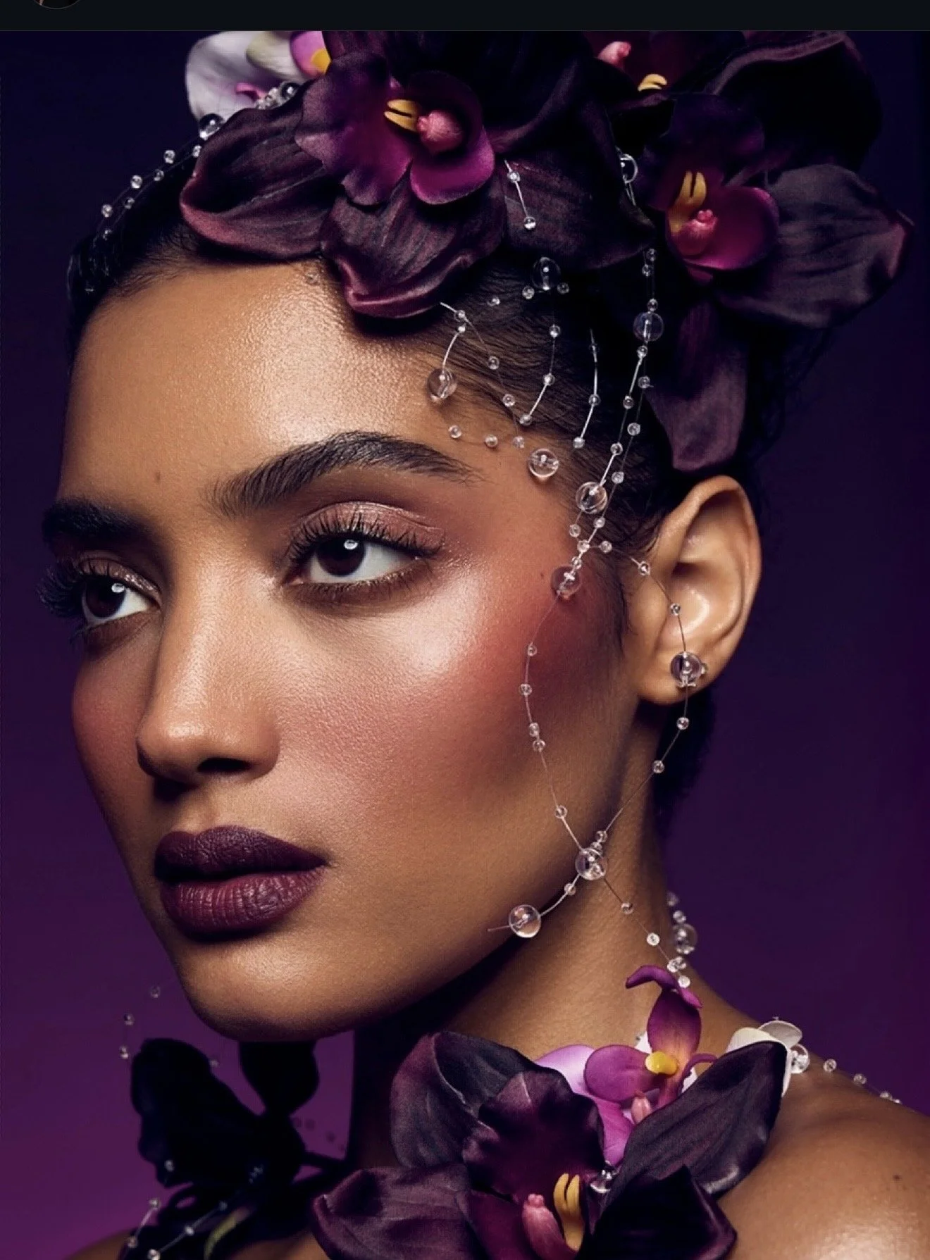 Close-up portrait of a woman with dark lipstick, eye makeup, and dark hair adorned with purple flowers and decorative beads and crystals.