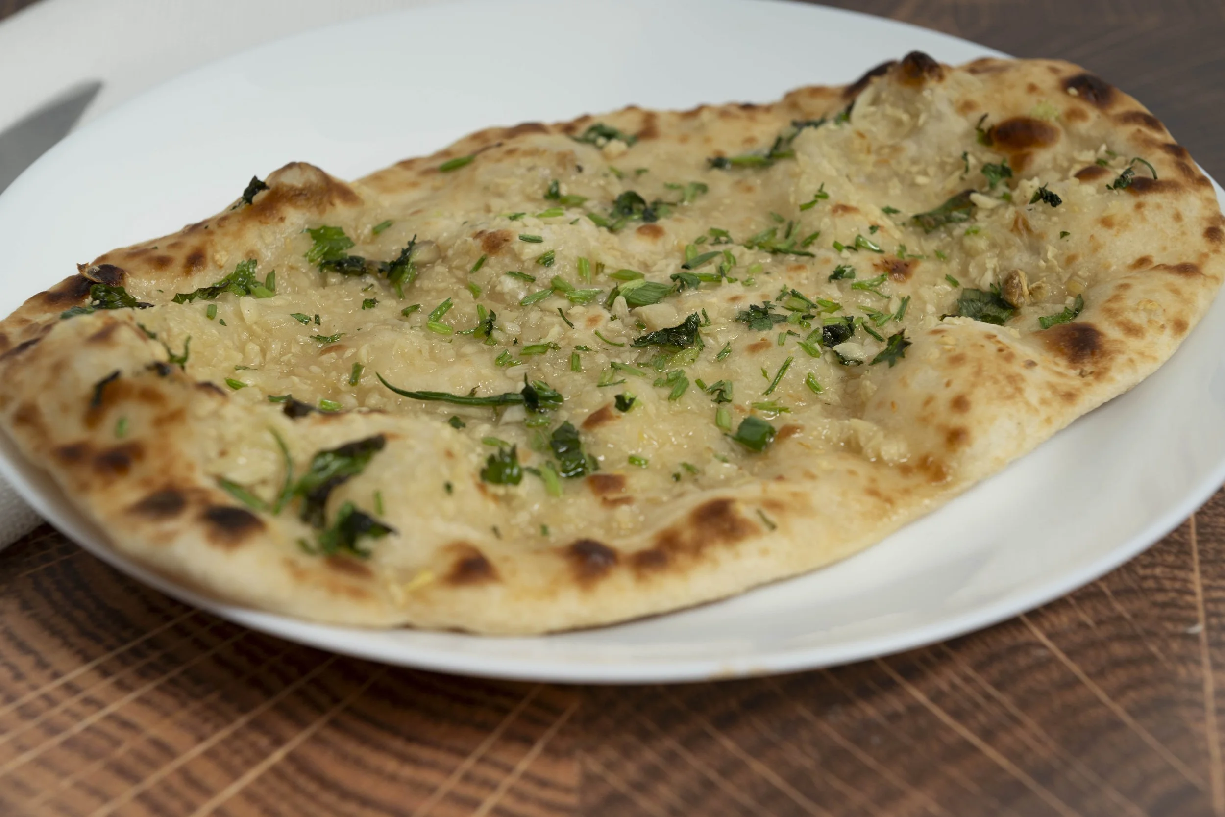 Close-up of a white plate with a piece of garlic naan topped with chopped herbs, placed on a wooden surface.