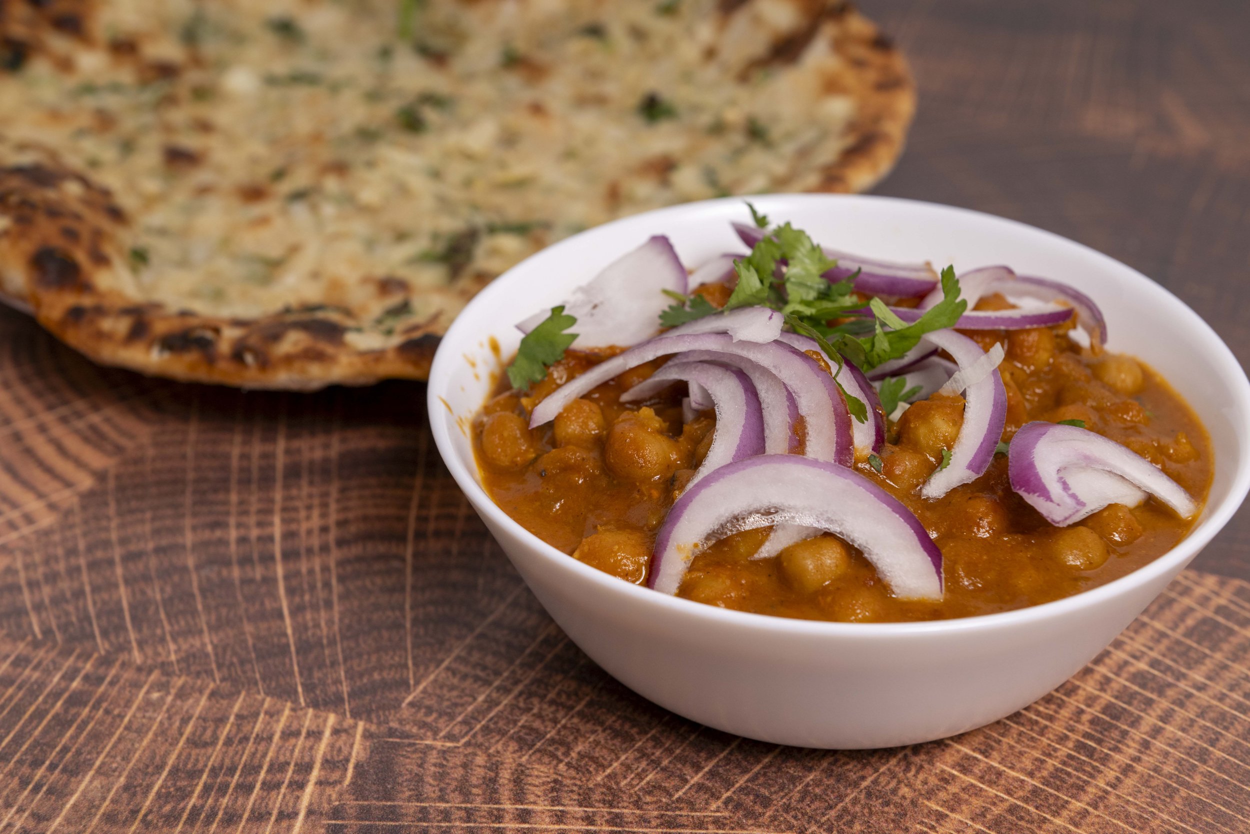 A white bowl of chickpea curry garnished with sliced red onions and cilantro, with naan bread in the background on a wooden surface.